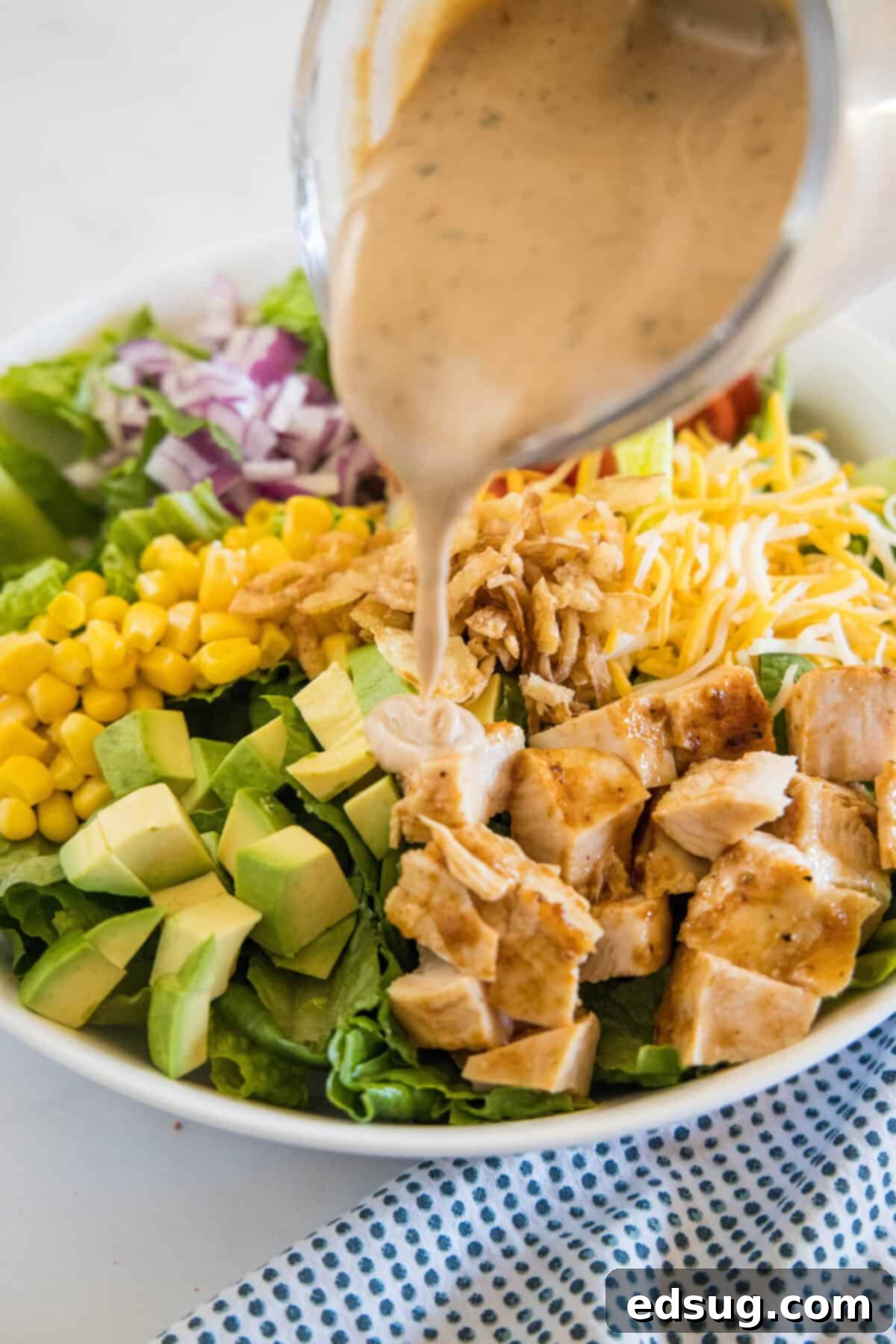 This BBQ chicken salad has juicy bites of sweet-smoky chicken tossed with tomatoes, avocado, and sweet corn in creamy barbecue ranch dressing. A spoon drizzling creamy barbecue ranch dressing generously over a vibrant BBQ chicken salad, ready to be tossed.