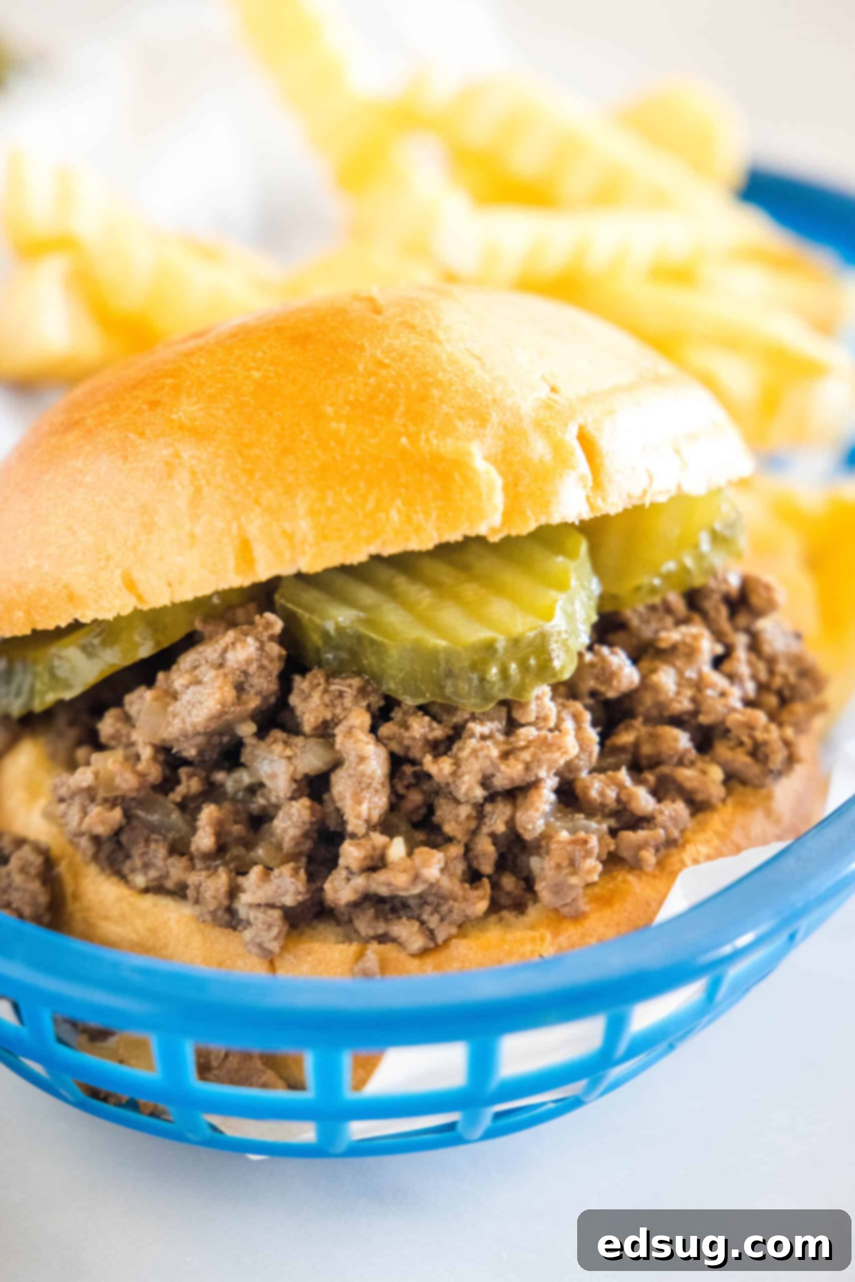 Discover how to make this authentic Maid-Rite loose meat sandwich recipe at home, a true Midwestern comfort food experience. A Maid Rite sandwich garnished with pickles in a blue plastic serving basket, with fries in the background.