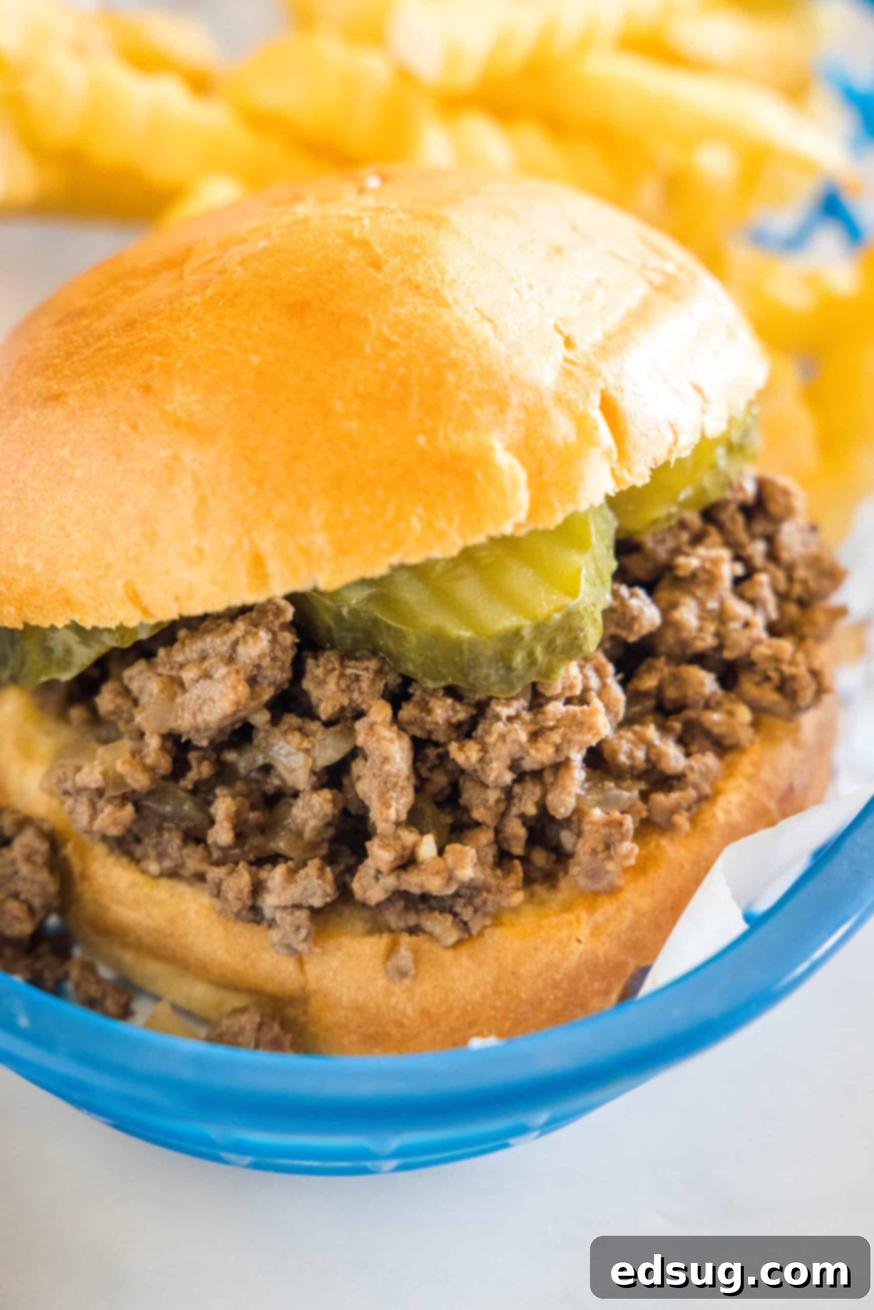Serve your delicious homemade Maid-Rite loose meat sandwich with classic sides like fries and pickles. A Maid Rite sandwich garnished with pickles in a blue plastic serving basket, next to a side of fries.