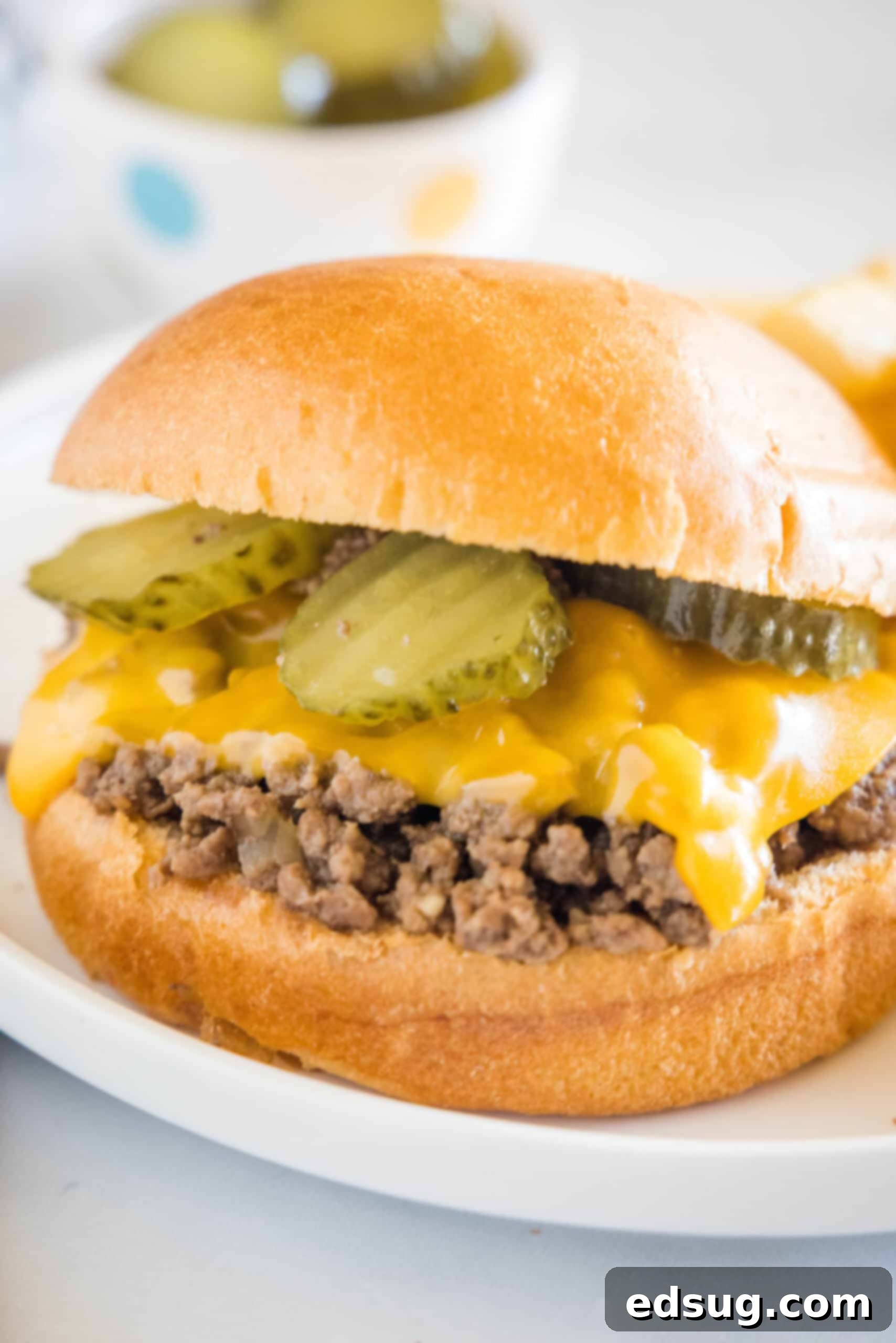 Enjoy a Maid-Rite loose meat sandwich with melted cheese and pickles on a white plate. A Maid Rite sandwich garnished with pickles and melted cheese on a white plate.