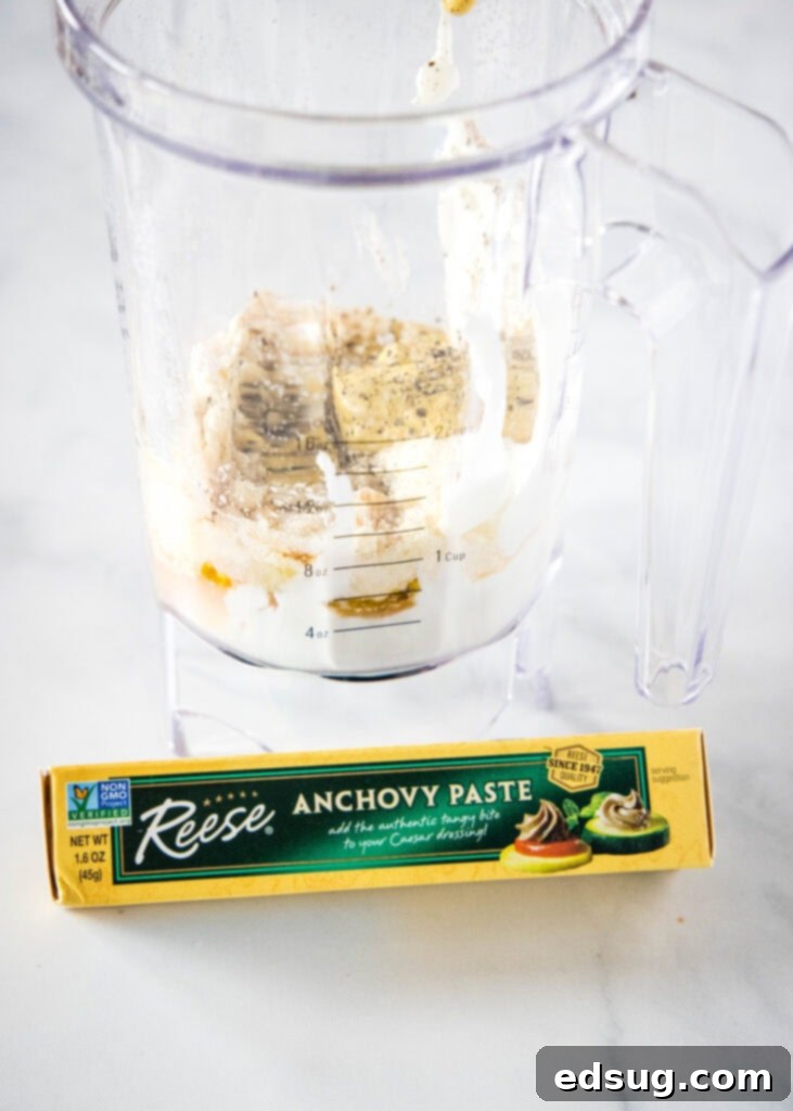 Signature Grilled Chicken Caesar 4 Caesar dressing ingredients in a blender, with a tube of anchovy paste positioned in front, highlighting the key components.