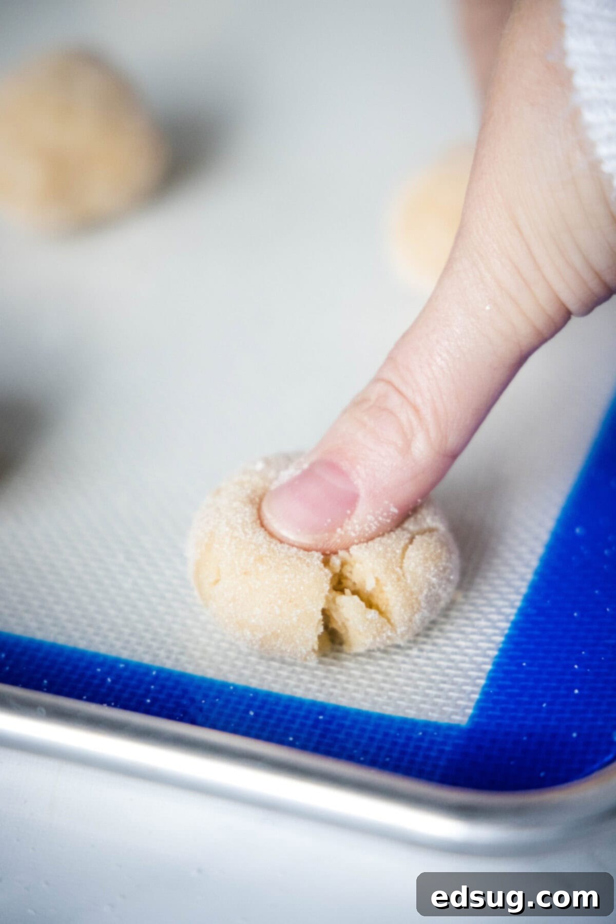 A baker's hand gently pressing a thumbprint into a perfectly rounded, raw Angel Crisp cookie dough ball on a baking sheet, preparing it for the oven.
