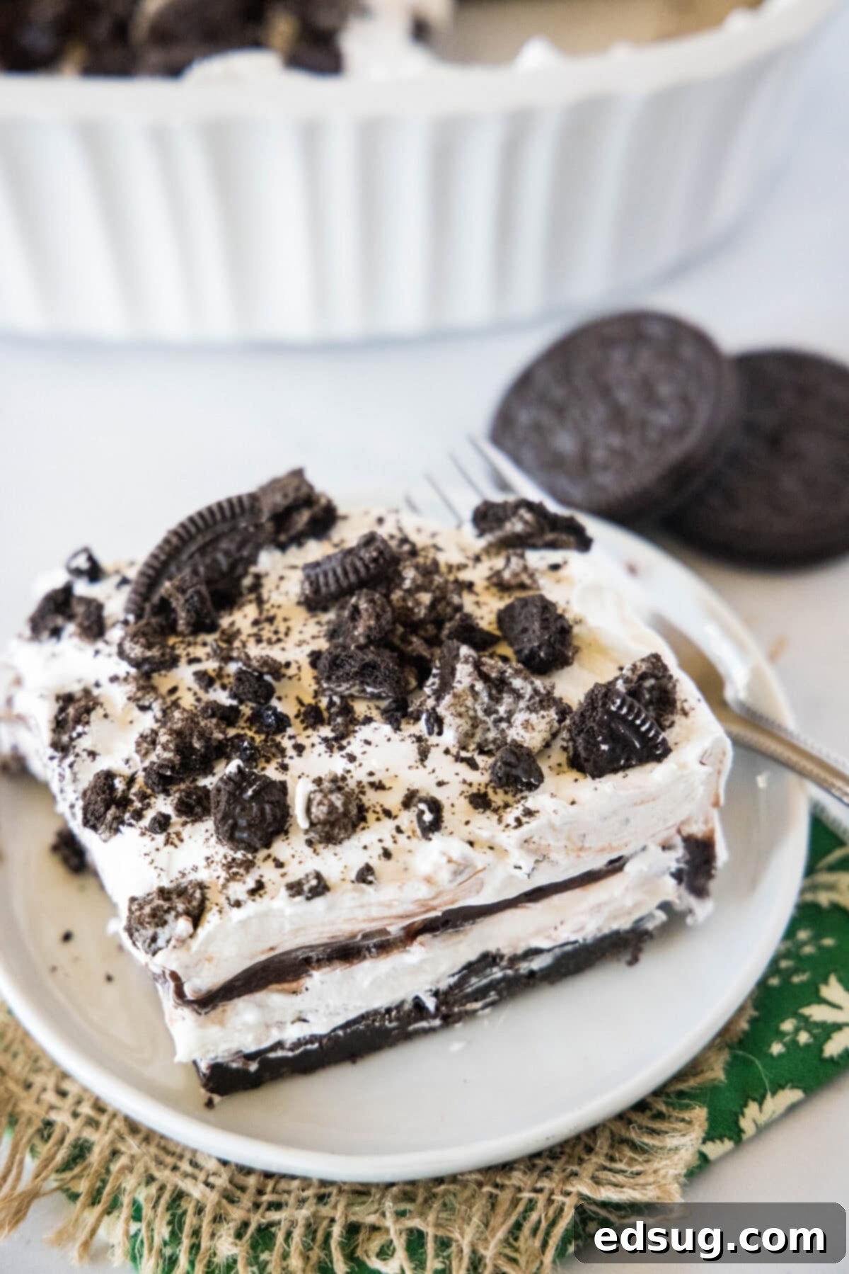 Overhead view of a slice of Oreo squares on a plate, topped with crumbled Oreos, next to a few Oreos, showcasing the distinct layers of crust, cream cheese, pudding, and whipped topping.