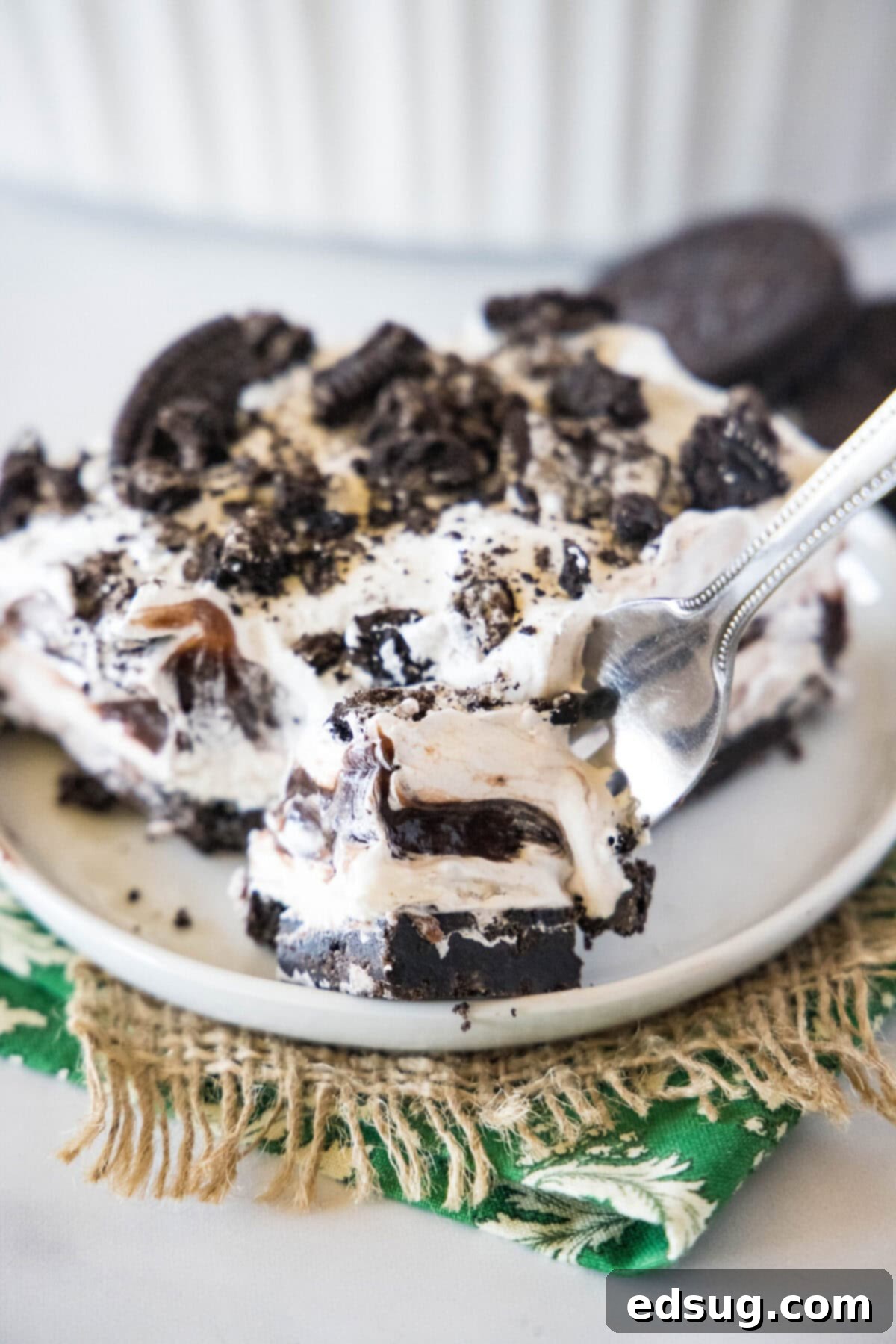 A fork holding a bite of layered no-bake Oreo lasagna, showing a perfect cross-section of the crust, cream cheese, pudding, and whipped topping, with a full slice on a plate blurred in the background.