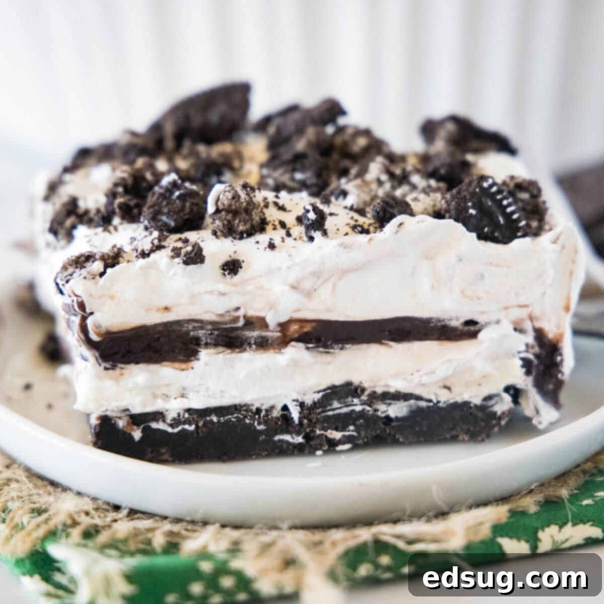 Close up of a perfectly sliced piece of no-bake Oreo lasagna on a white plate, showing the distinct dark cookie crust, white cream cheese layer, brown pudding layer, and a final topping of whipped cream and Oreo crumbles.
