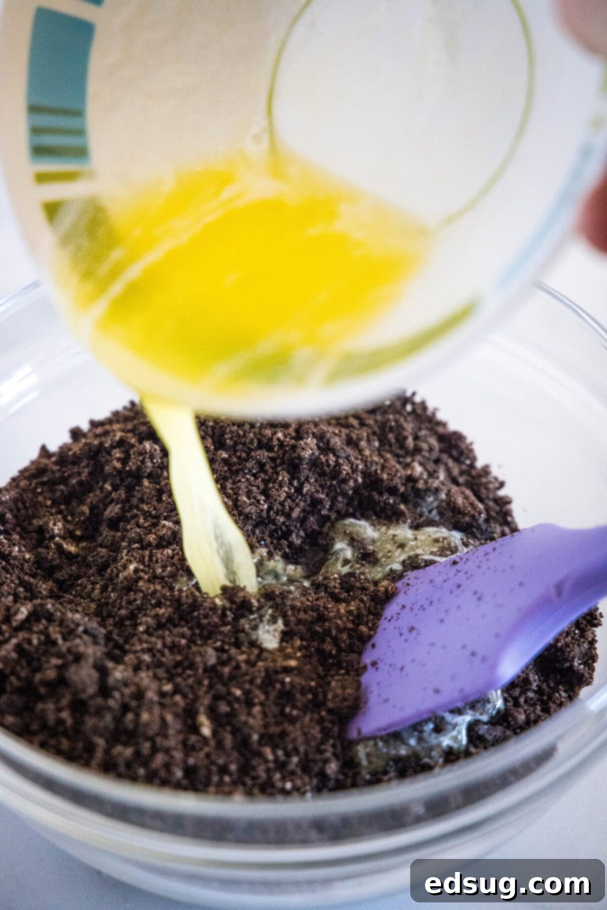 A glass measuring cup pouring melted butter into a bowl filled with finely crushed Oreo crumbs, next to a rubber spatula, ready to be mixed for the crust of the no-bake dessert.