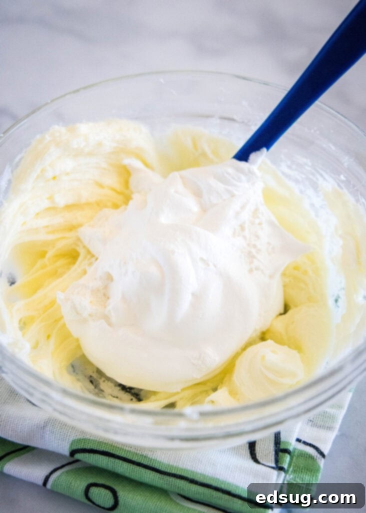 A rubber spatula gently folding a portion of Cool Whip into the beaten cream cheese mixture in a large mixing bowl, ensuring a light and airy texture.