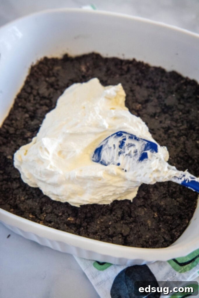 A rubber spatula carefully spreading the white, fluffy cream cheese mixture over the dark, firm Oreo cookie crust in a glass baking dish, forming the first creamy layer.
