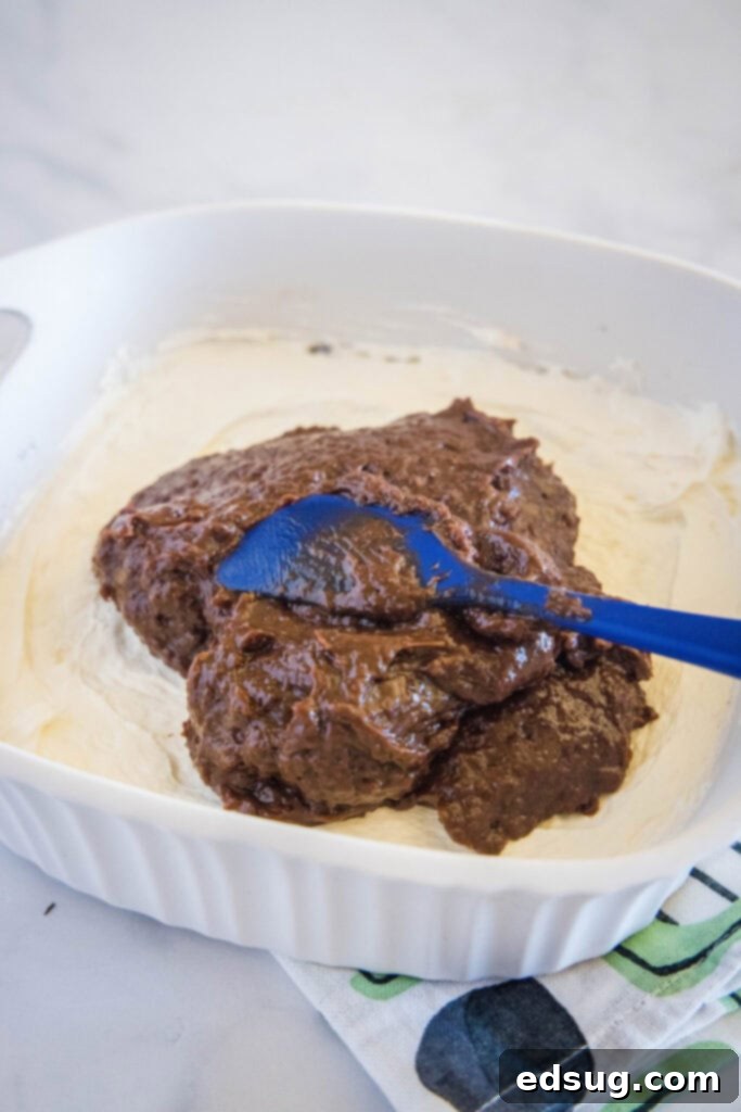 A rubber spatula gently spreading rich chocolate pudding over the white cream cheese layer in a baking dish, creating the second distinct layer of the no-bake dessert.