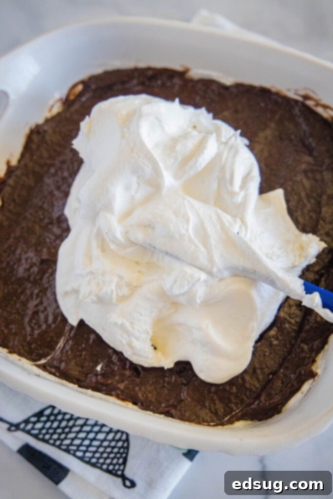 A spatula gently spreading a generous layer of white Cool Whip over the brown chocolate pudding layer in a baking dish, preparing the final creamy layer of the Oreo lasagna.