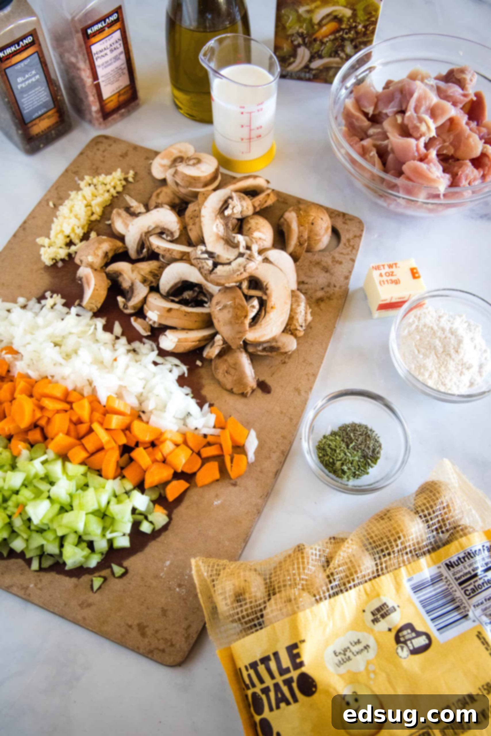 Various raw ingredients for chicken stew laid out on a cutting board, including chicken thighs, carrots, celery, onion, garlic, and mushrooms.