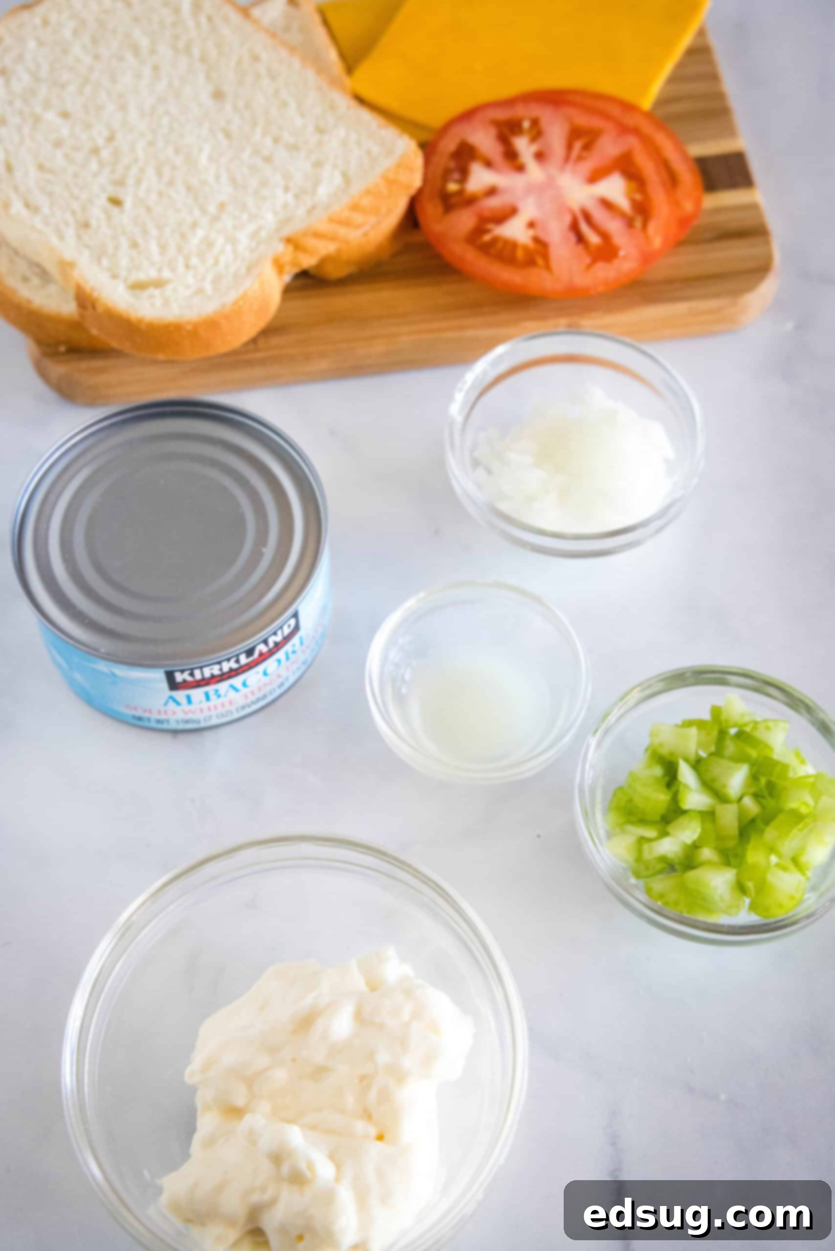 Fresh ingredients for making the best tuna melt, including a can of tuna, mayonnaise, celery, onion, lemon, bread, tomatoes, and cheese, neatly arranged on a wooden board.