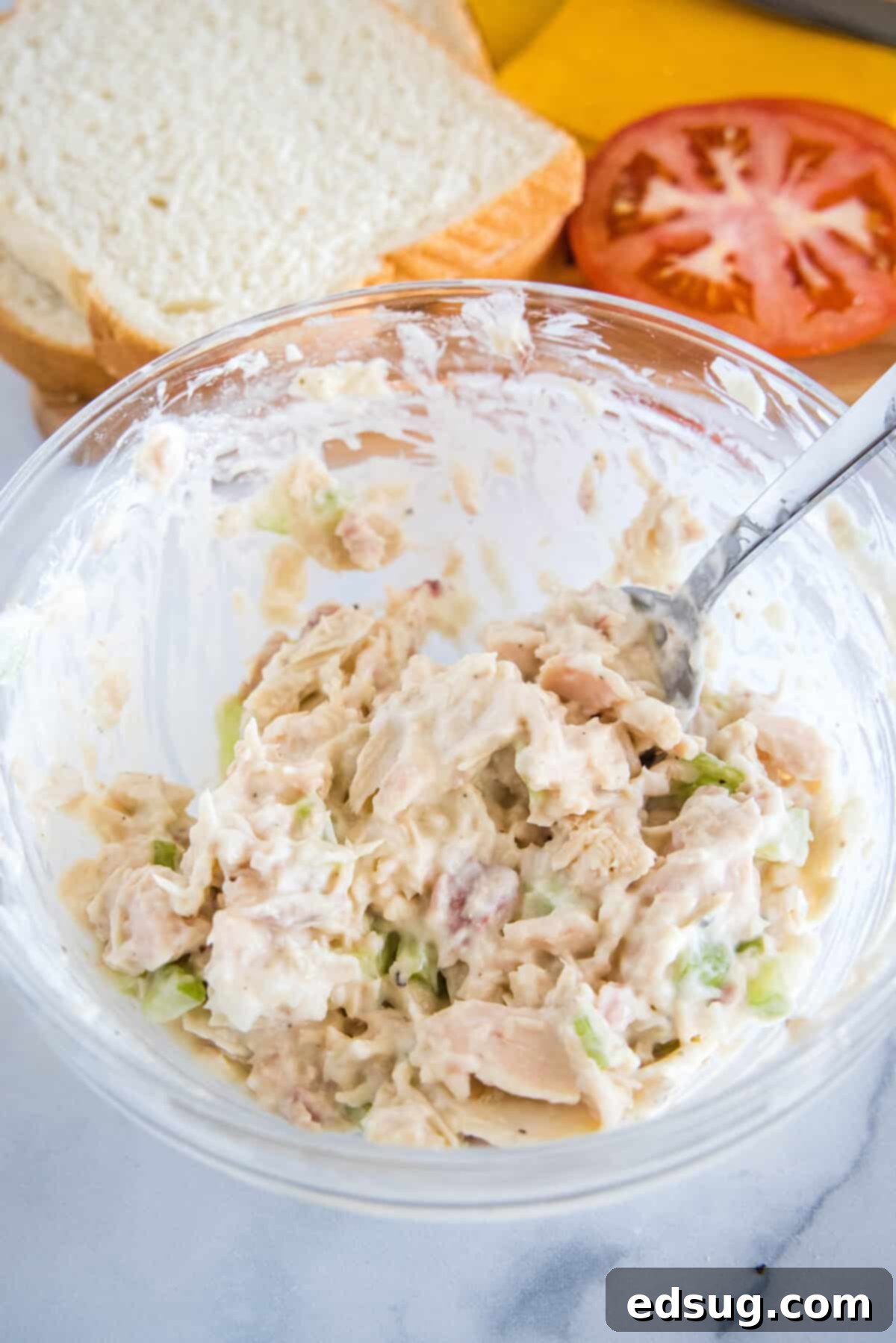 A close-up of the mixed tuna salad in a glass bowl with a fork, alongside slices of fresh bread and ripe tomatoes in the background, ready for sandwich assembly.