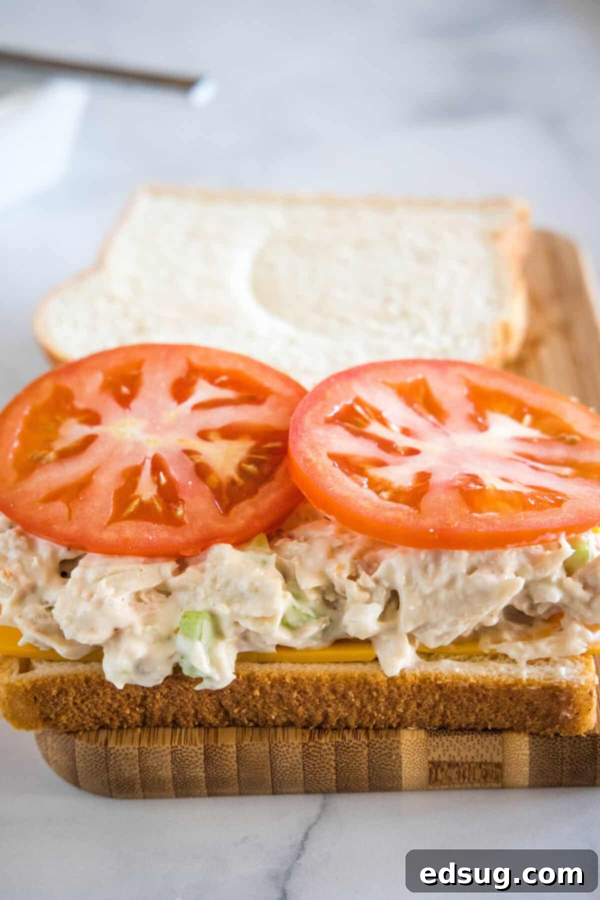 An open-faced tuna melt sandwich assembled on a wooden cutting board, with tuna salad, sliced tomatoes, and cheese layered on buttered bread, ready to be grilled.