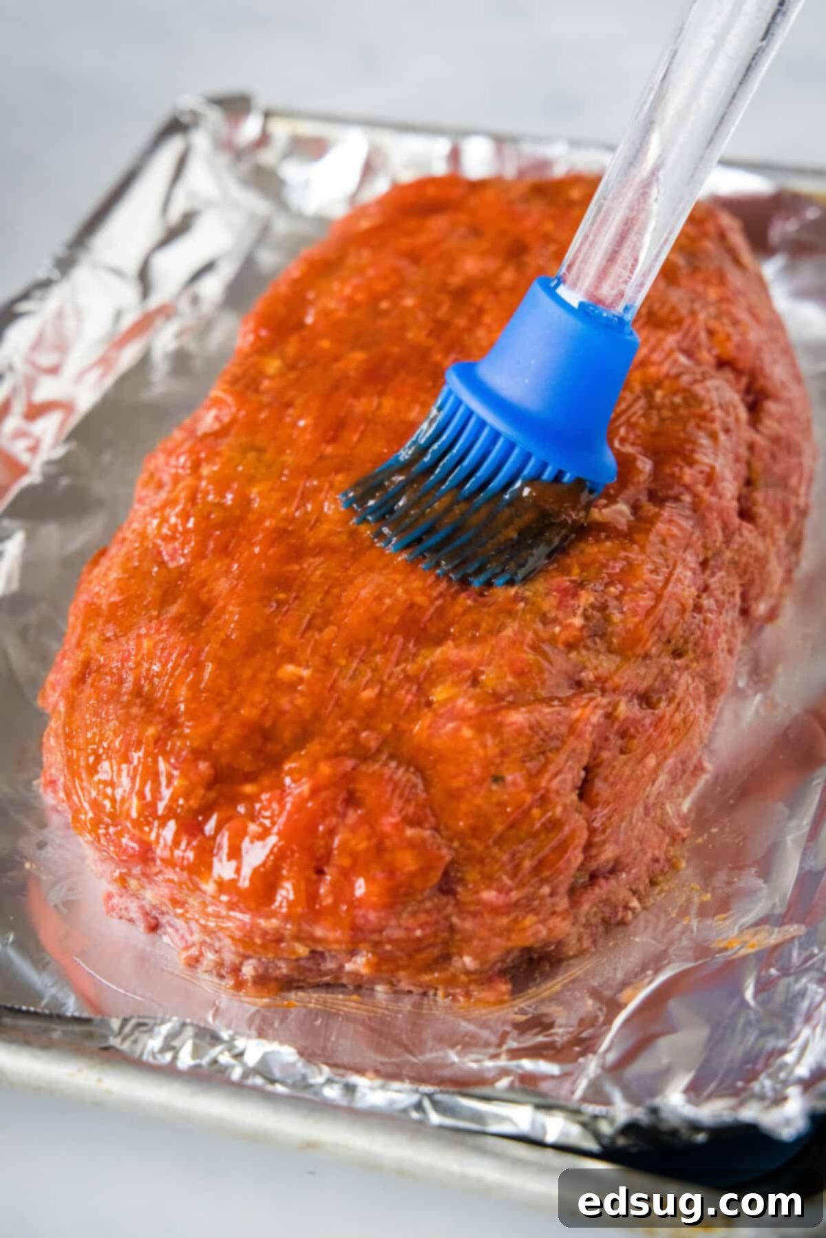 A blue basting brush brushing sauce over the outside of onion soup meatloaf on a lined baking sheet. A blue basting brush brushing sauce over the outside of onion soup meatloaf on a lined baking sheet.