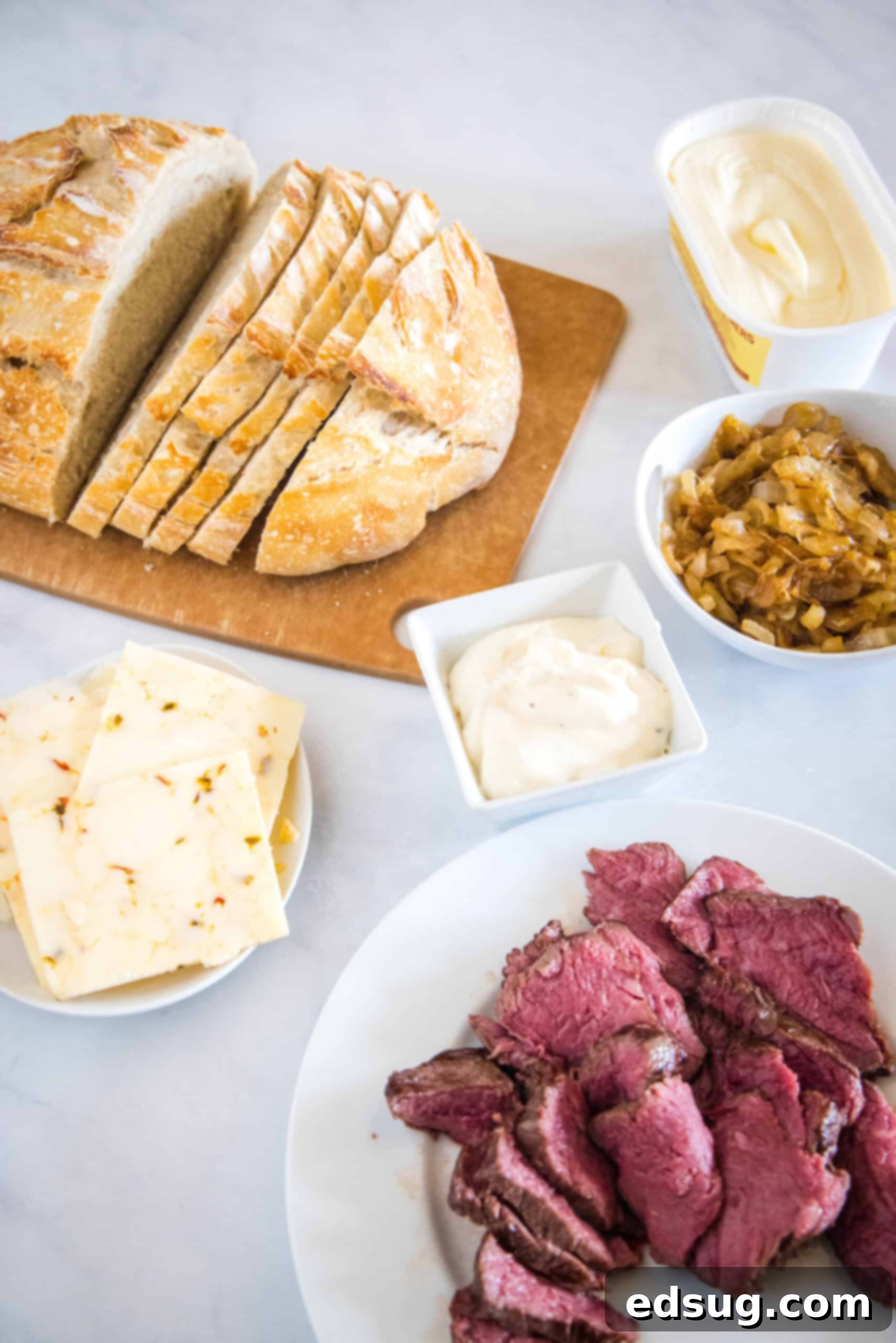 A vibrant display of fresh steak sandwich ingredients, including sirloin, sourdough bread, cheese, caramelized onions, garlic aioli, and butter.