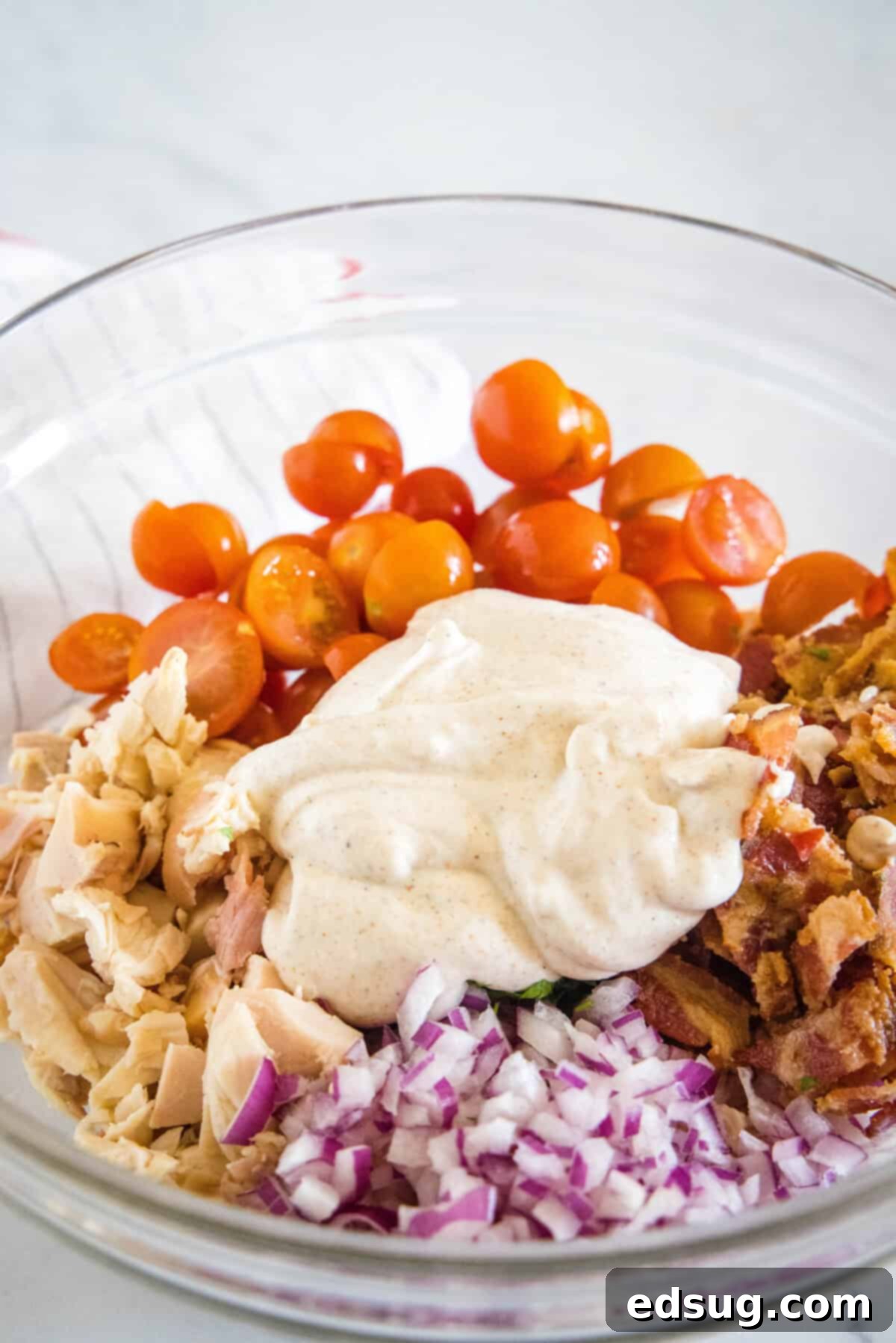 This BLT chicken salad is loaded with juicy shredded chicken, bacon, cherry tomatoes, and crispy red onion tossed with creamy mayo dressing. Dressing added to BLT chicken salad ingredients in a glass bowl.