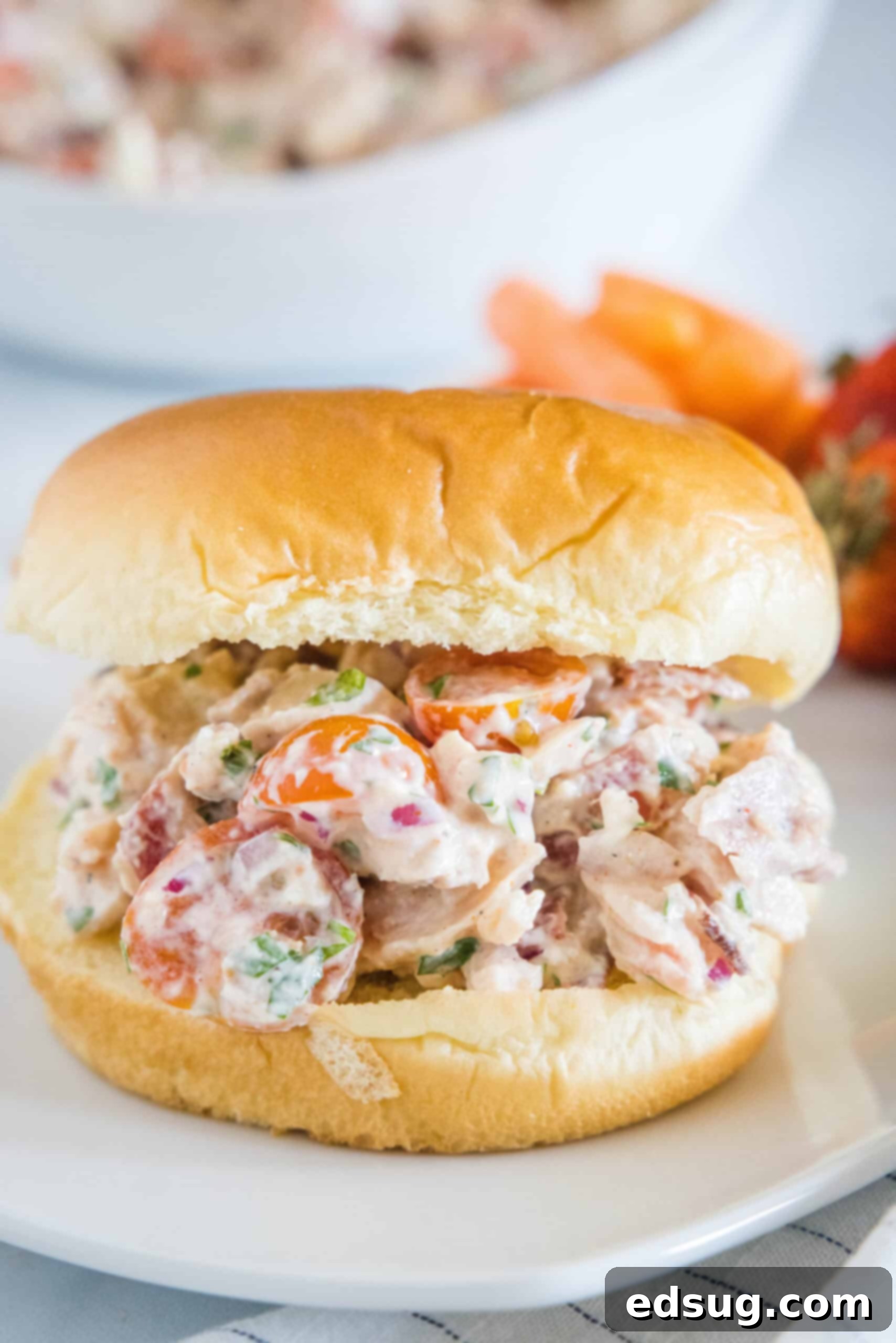 This BLT chicken salad is loaded with juicy shredded chicken, bacon, cherry tomatoes, and crispy red onion tossed with creamy mayo dressing. A BLT chicken salad sandwich on a white plate with a side of carrot sticks and fresh strawberries in the background.