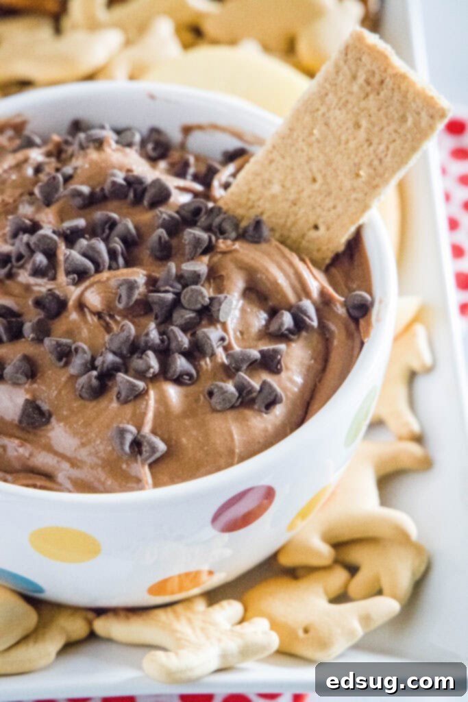 A graham cracker being dipped into a bowl of creamy, chocolatey brownie batter dip, showcasing its rich texture and inviting appearance.