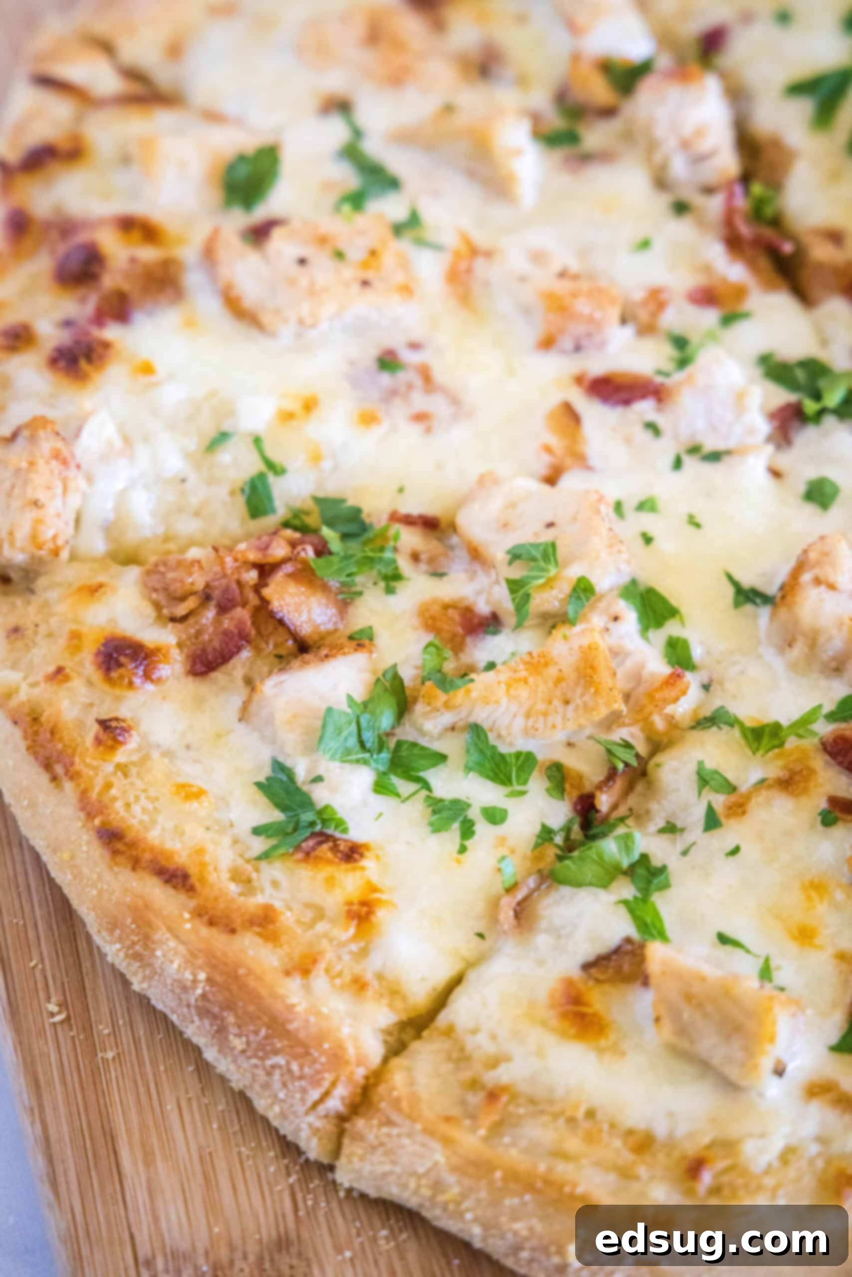 Close up of a chicken alfredo pizza cut into slices on a wooden butting board, showcasing the creamy sauce, chicken, and bacon toppings.
