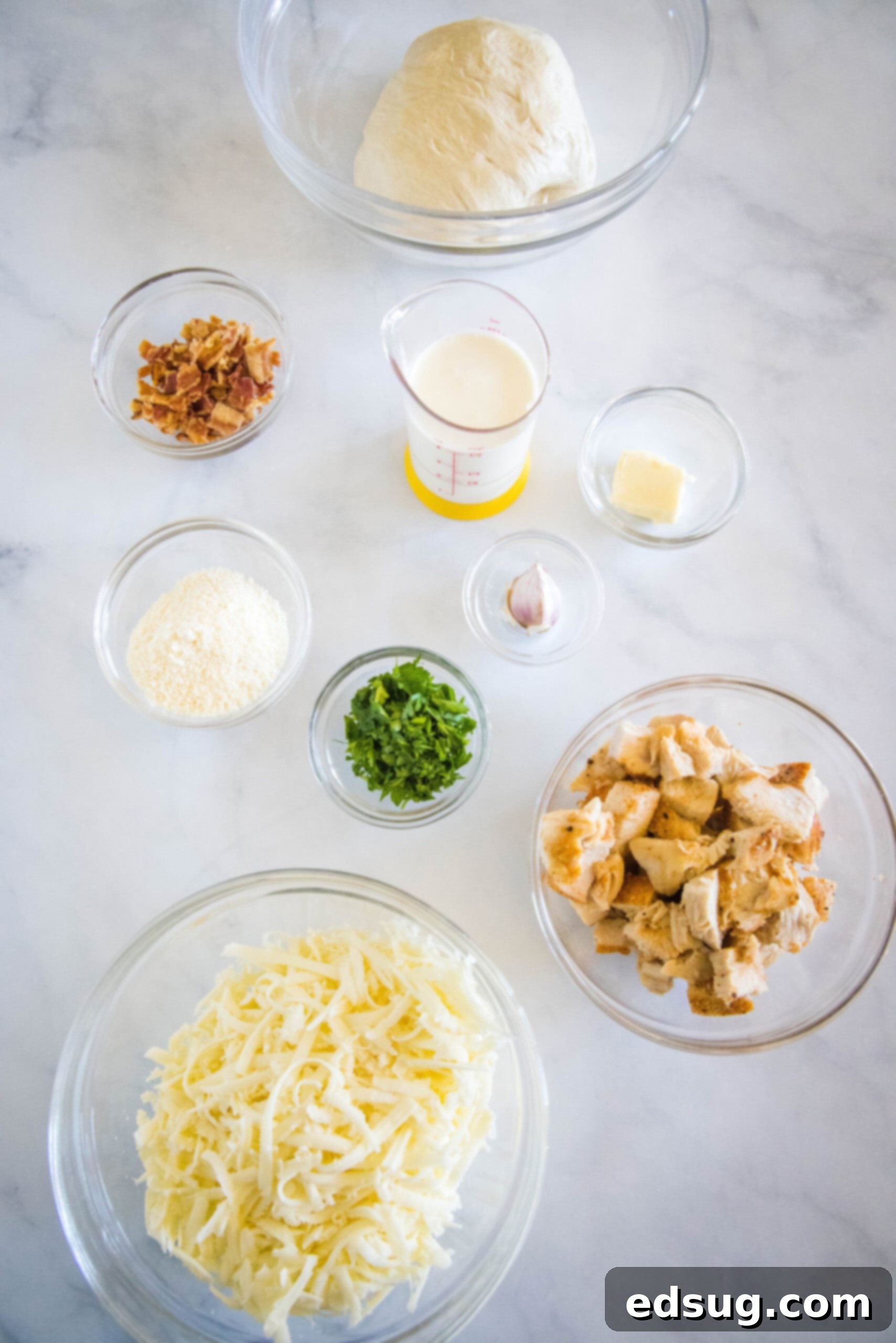 Chicken alfredo pizza ingredients laid out on a table, including dough, sauce, cheese, chicken, and bacon.