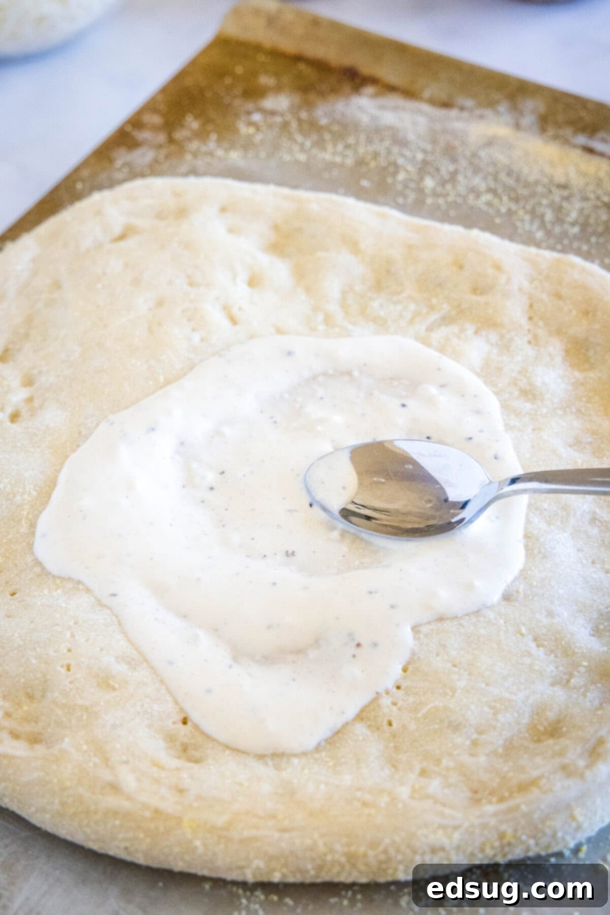 A spoon spreading creamy alfredo sauce evenly over a parbaked pizza crust.