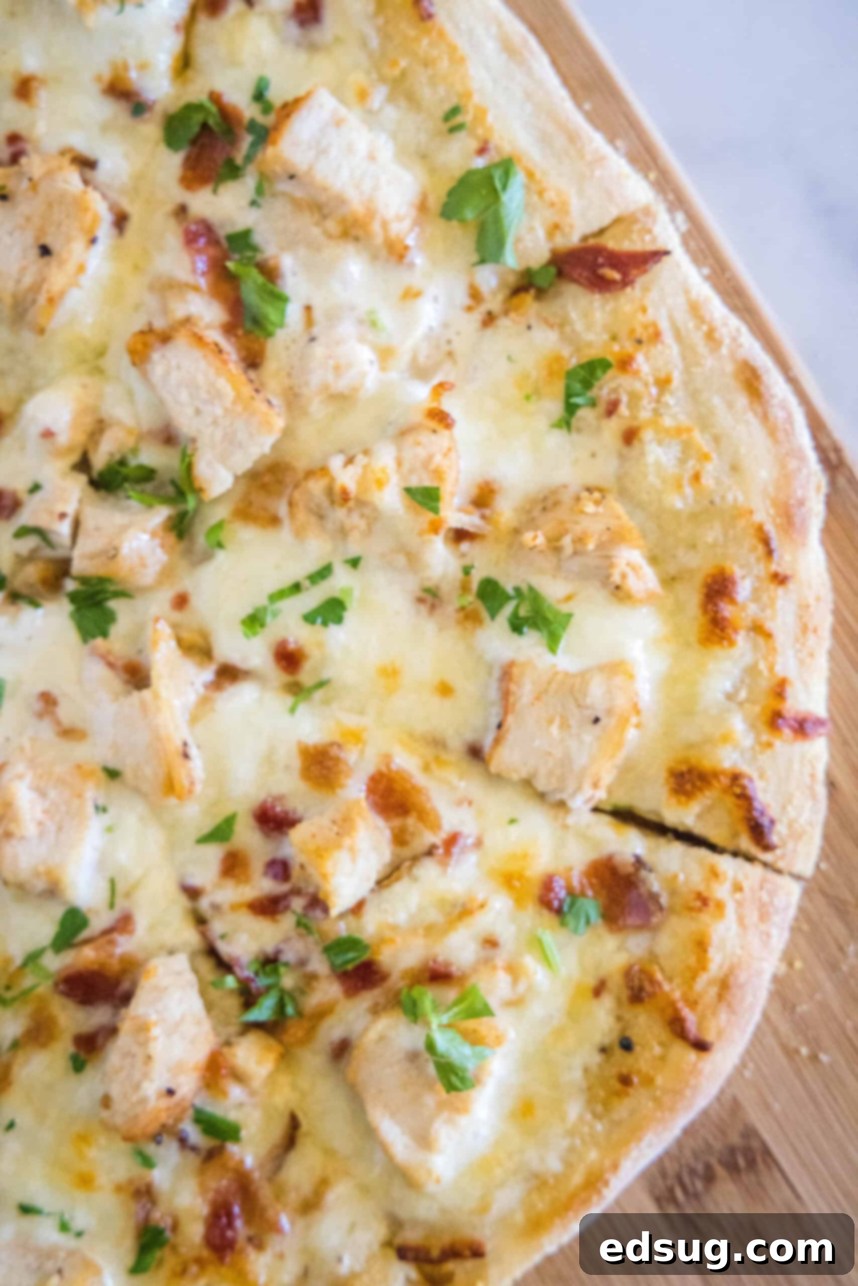 Overhead view of a cooked chicken alfredo pizza cut into slices on a wooden cutting board, garnished with fresh parsley.