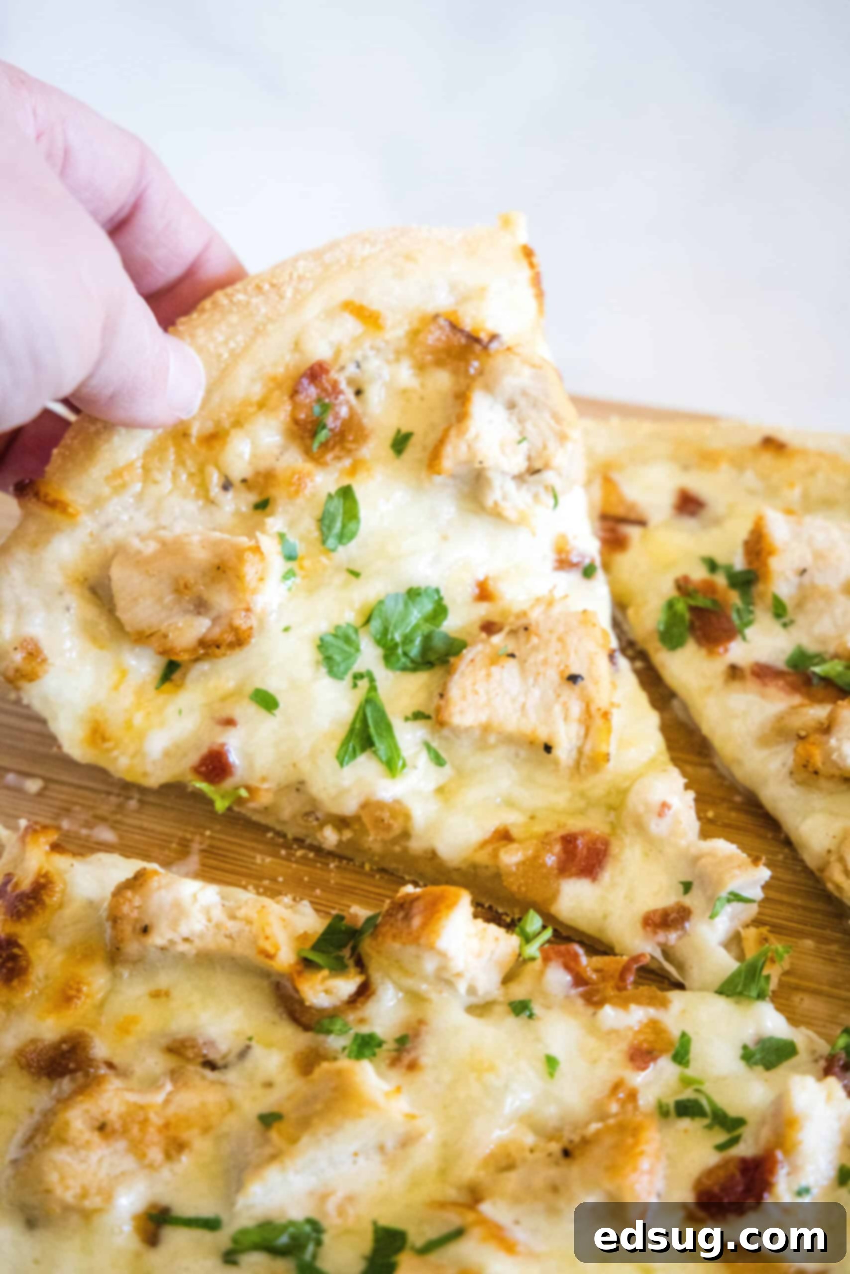 A hand lifting a single slice of chicken alfredo pizza, showing the melted cheese and toppings, from a wooden cutting board.