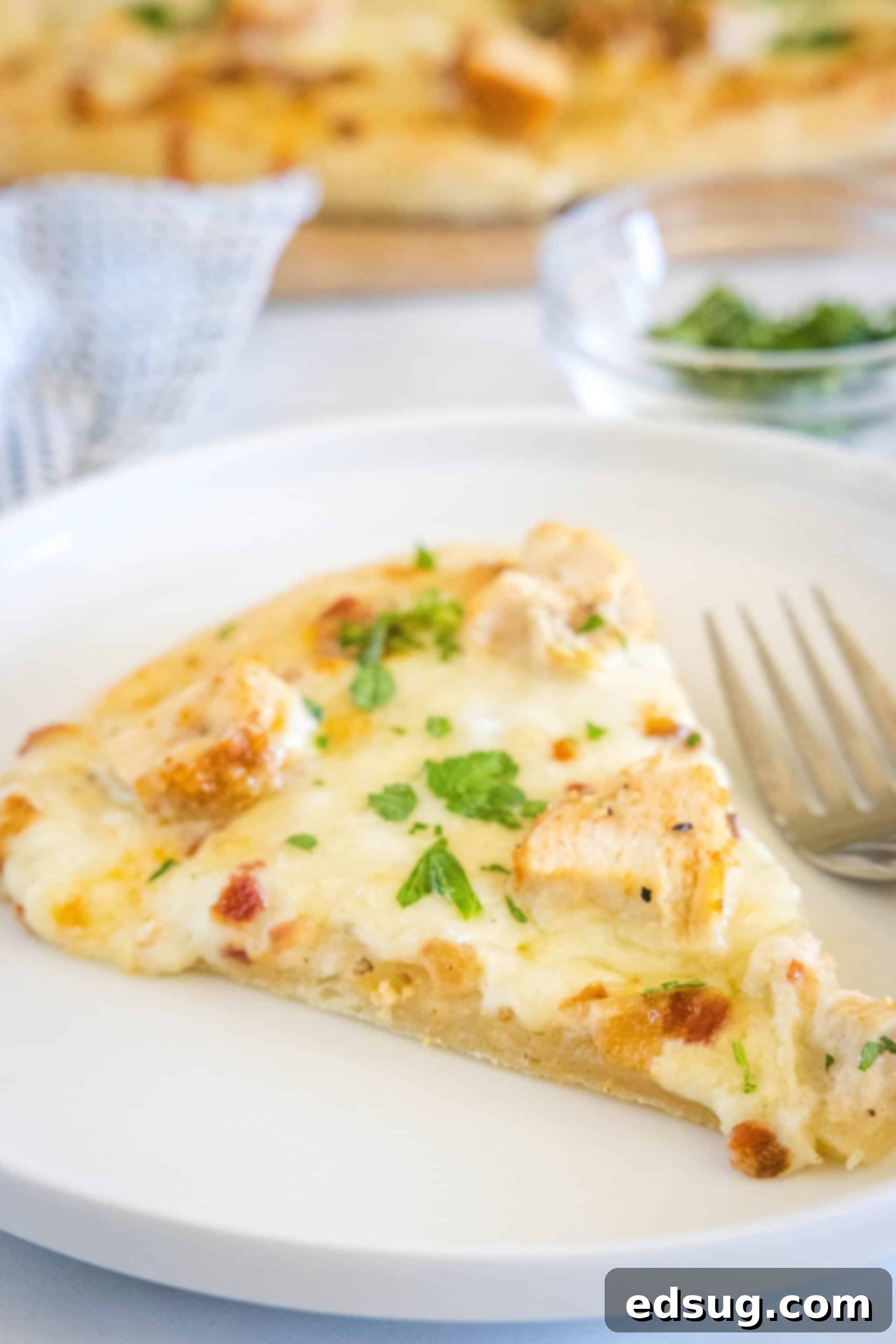 A single slice of chicken alfredo pizza on a white plate next to a fork, ready to be eaten.