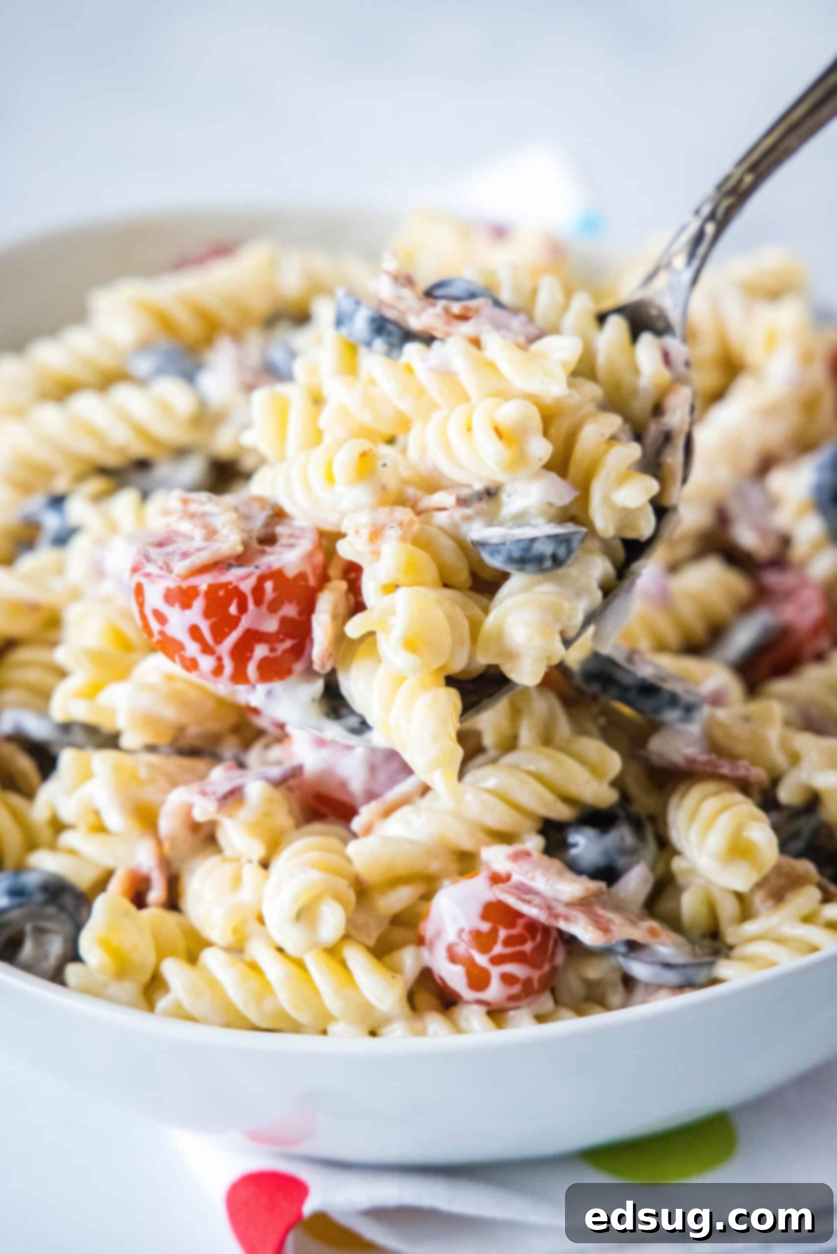 A large serving spoon scooping ranch pasta salad from a white bowl, showcasing crispy bacon, tomatoes, olives, and rotini pasta.