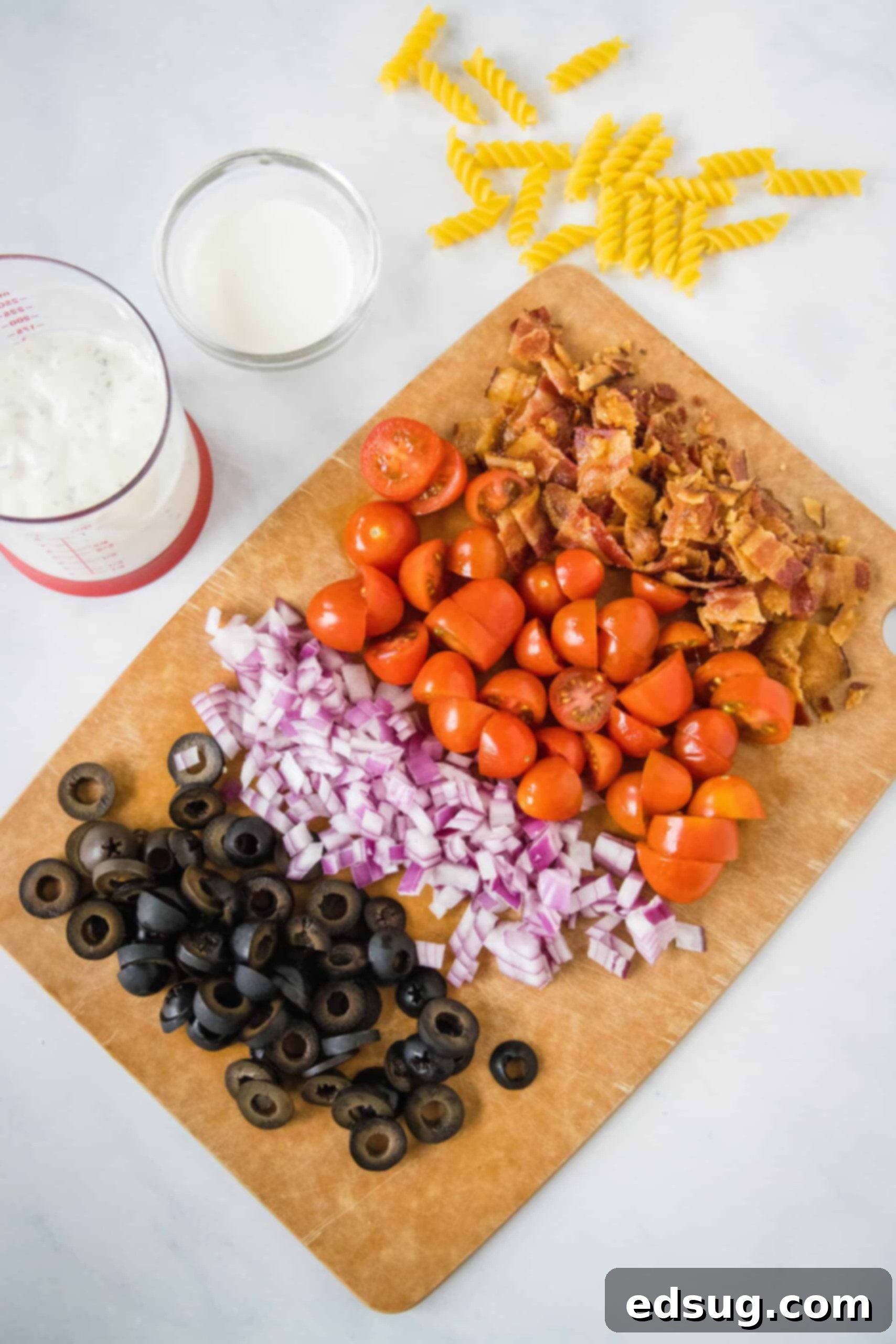 Ranch pasta salad ingredients including uncooked pasta, chopped onion, tomatoes, olives, and crumbled bacon, prepared on a wooden cutting board.
