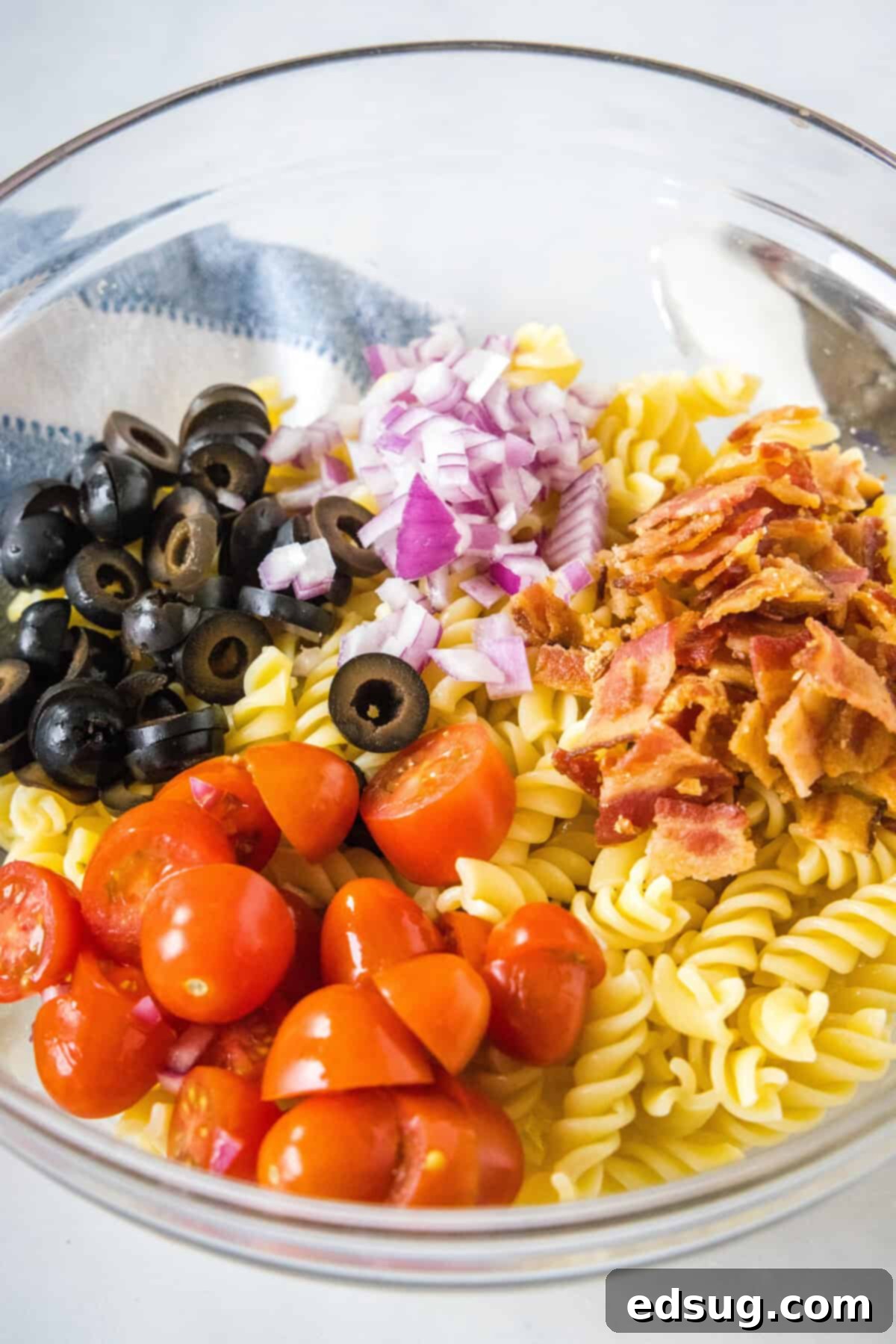 Ranch pasta salad ingredients including cooked pasta, tomatoes, red onion, bacon, and black olives combined in a large glass bowl, ready for dressing.