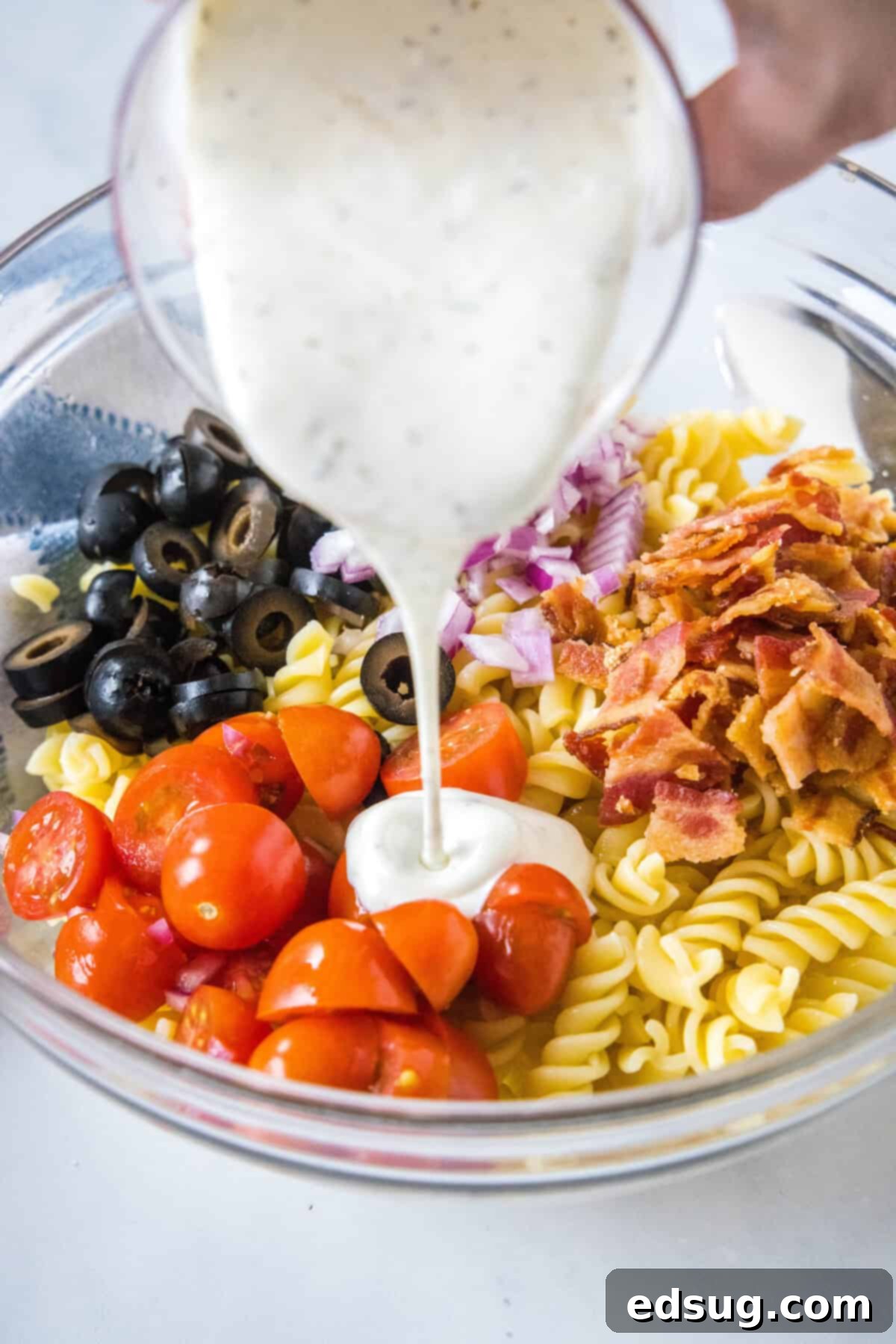 Creamy ranch dressing being poured generously over the combined pasta salad ingredients in a clear glass bowl, preparing for tossing.