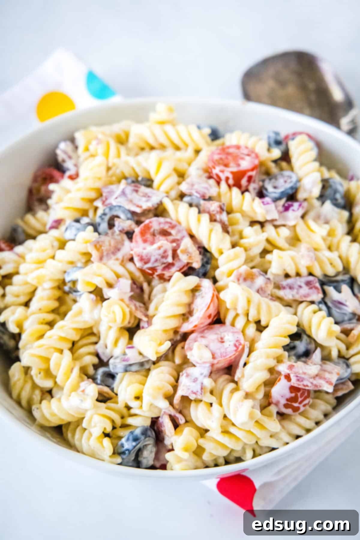 A serving spoon tossing the ranch pasta salad in a white bowl, showing all ingredients thoroughly coated in the creamy dressing.