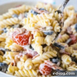 A large serving spoon scooping ranch pasta salad from a white bowl, with crisp bacon and fresh vegetables visible.