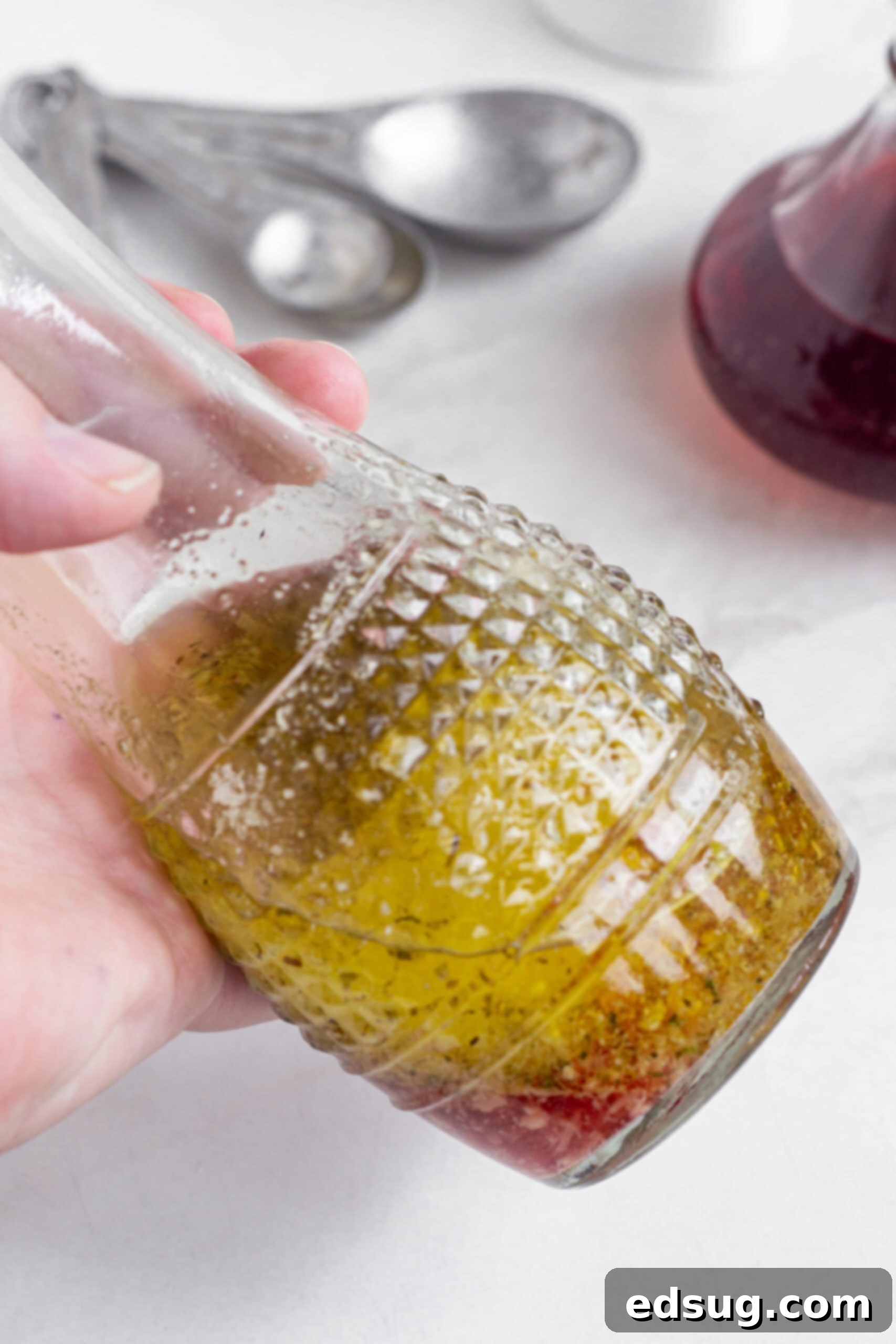 A hand holding up a glass carafe of red wine vinaigrette against a rustic background, highlighting its rich color.