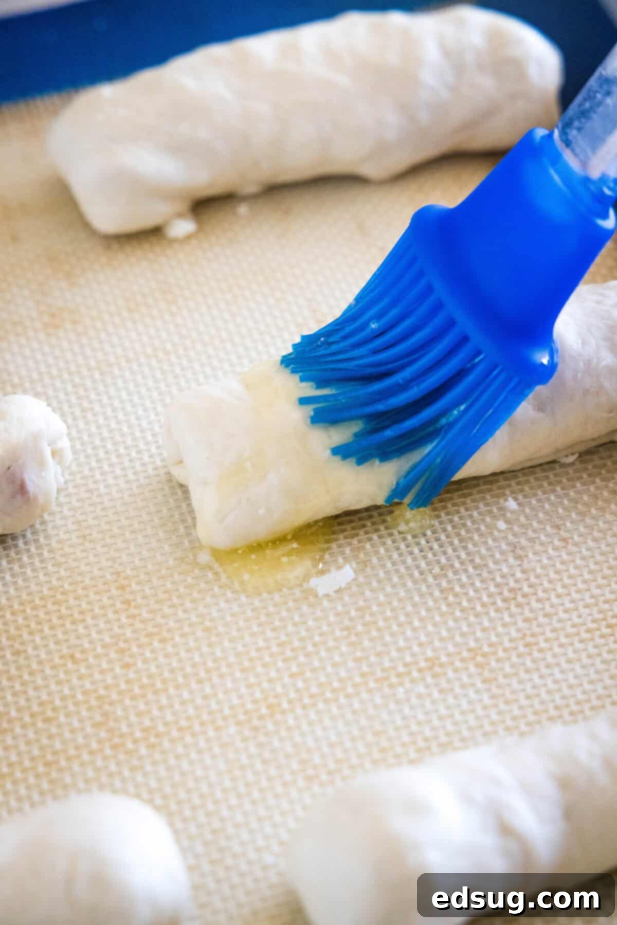 These pepperoni rolls are an easy snack made from soft, fluffy pizza dough filled with melty mozzarella cheese and juicy pepperoni slices. A blue rubber basting brush coating the top of an unbaked pepperoni roll with melted butter.