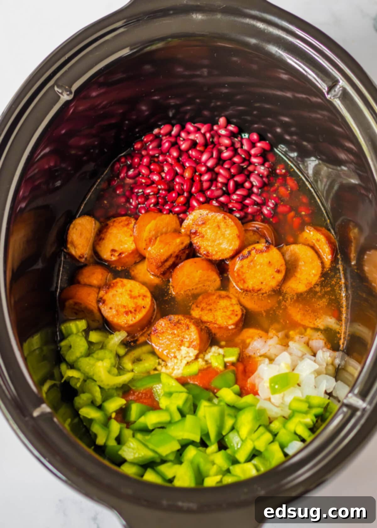 A crock pot filled with dried red beans, sliced sausage, celery, onions, green bell pepper, tomato sauce, and chicken broth.