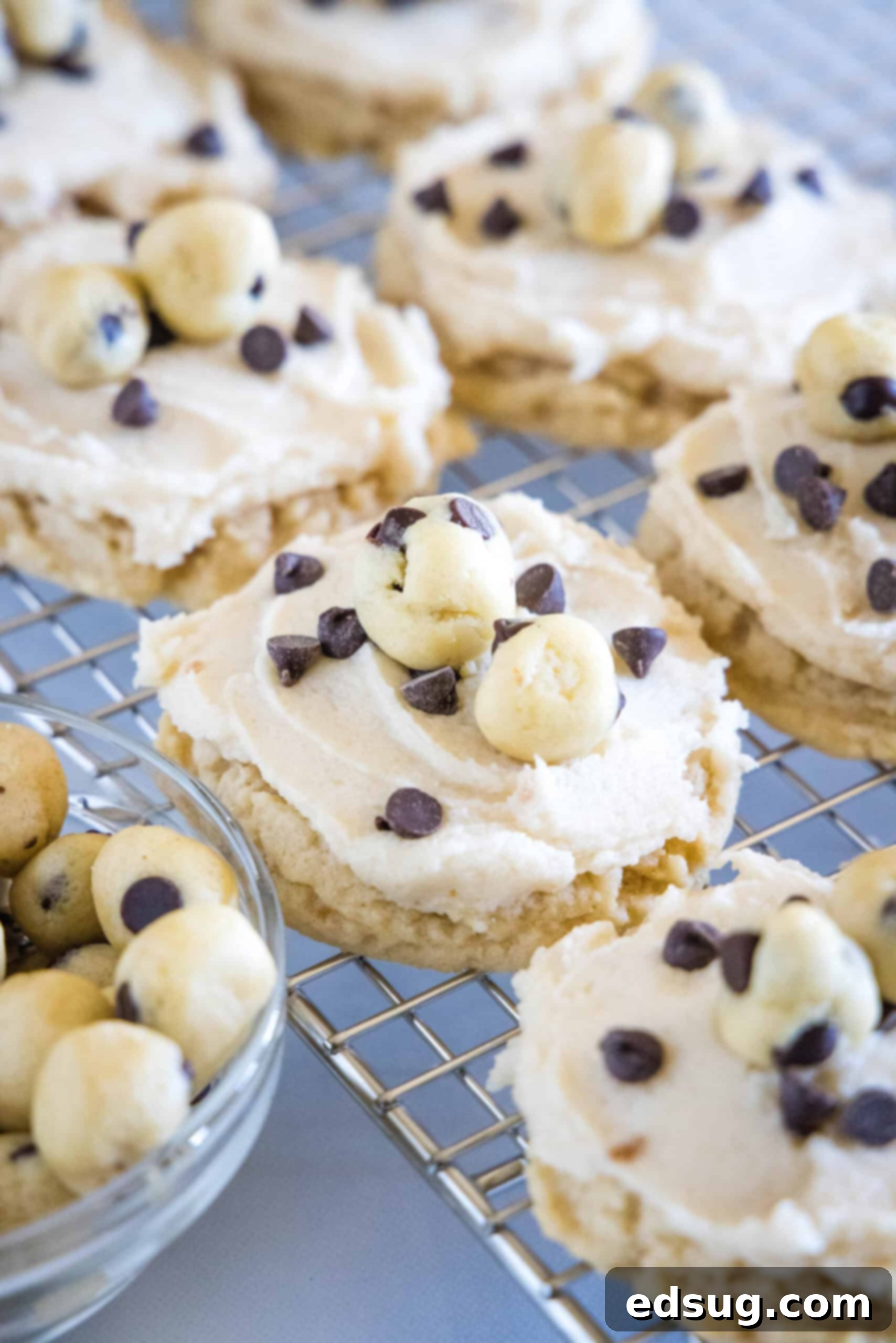 These cookie dough cookies are bakery-style sugar cookies topped with creamy cookie dough frosting, edible cookie dough, and chocolate chips! Beautifully frosted cookie dough cookies resting on a cooling wire rack, adorned with edible cookie dough pieces and mini chocolate chips.