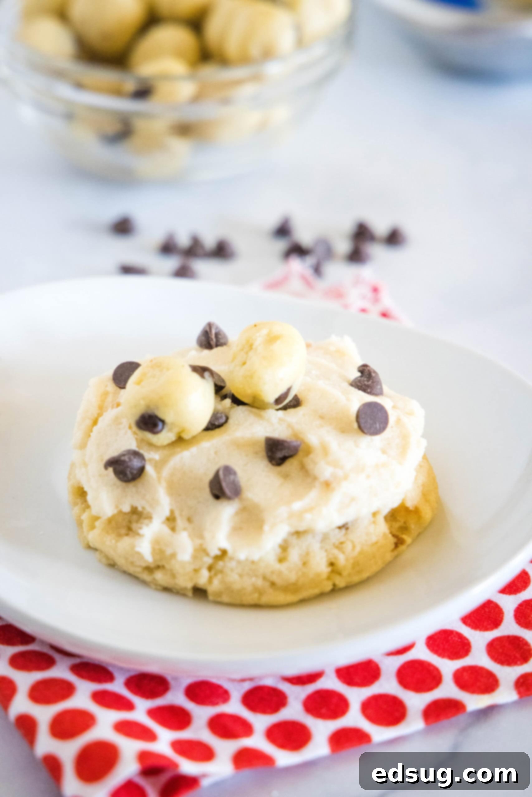 These cookie dough cookies are bakery-style sugar cookies topped with creamy cookie dough frosting, edible cookie dough, and chocolate chips! A single, perfectly frosted cookie dough cookie on a pristine white plate, showcasing its texture and toppings.
