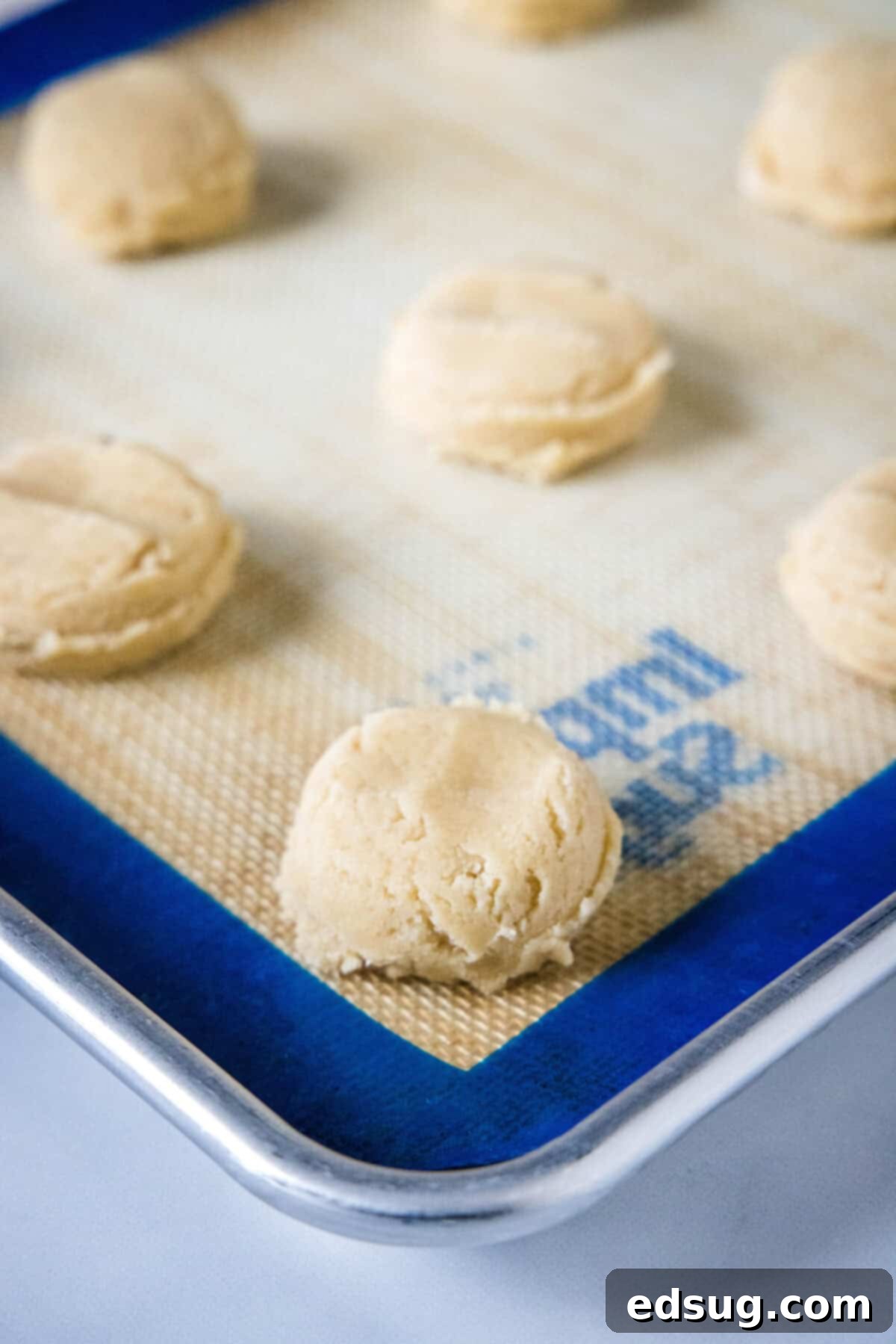 These cookie dough cookies are bakery-style sugar cookies topped with creamy cookie dough frosting, edible cookie dough, and chocolate chips! Portioned cookie dough balls arranged on a silpat-lined baking sheet, ready to be baked into delicious cookies.