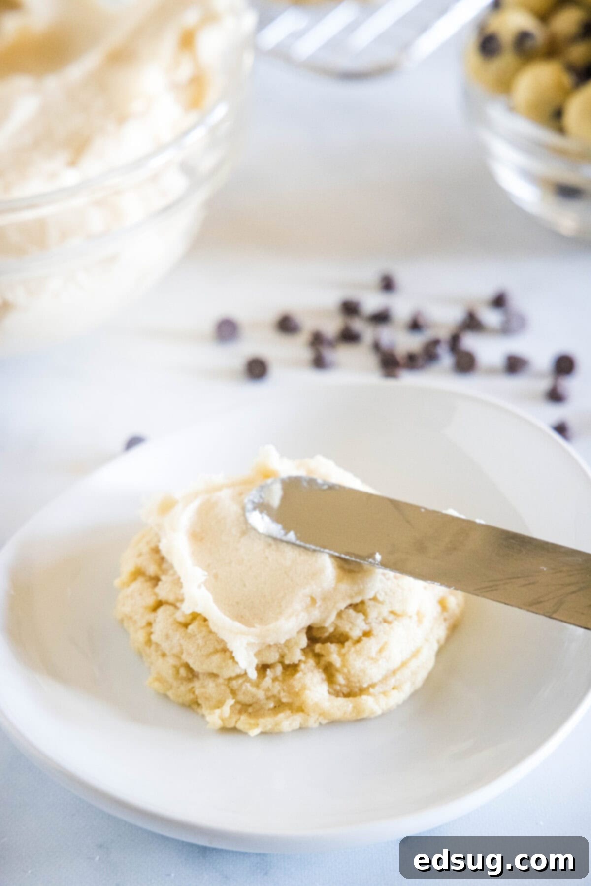 These cookie dough cookies are bakery-style sugar cookies topped with creamy cookie dough frosting, edible cookie dough, and chocolate chips! A smooth, creamy brown sugar frosting being expertly spread over a cooled sugar cookie with an offset spatula on a white plate.