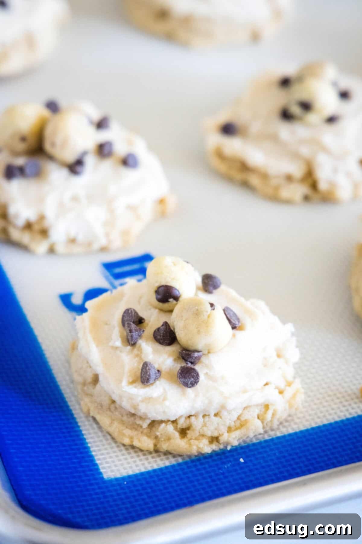 These cookie dough cookies are bakery-style sugar cookies topped with creamy cookie dough frosting, edible cookie dough, and chocolate chips! Finished cookie dough cookies, beautifully frosted and topped with mini edible cookie dough balls and chocolate chips, arranged on a baking sheet.