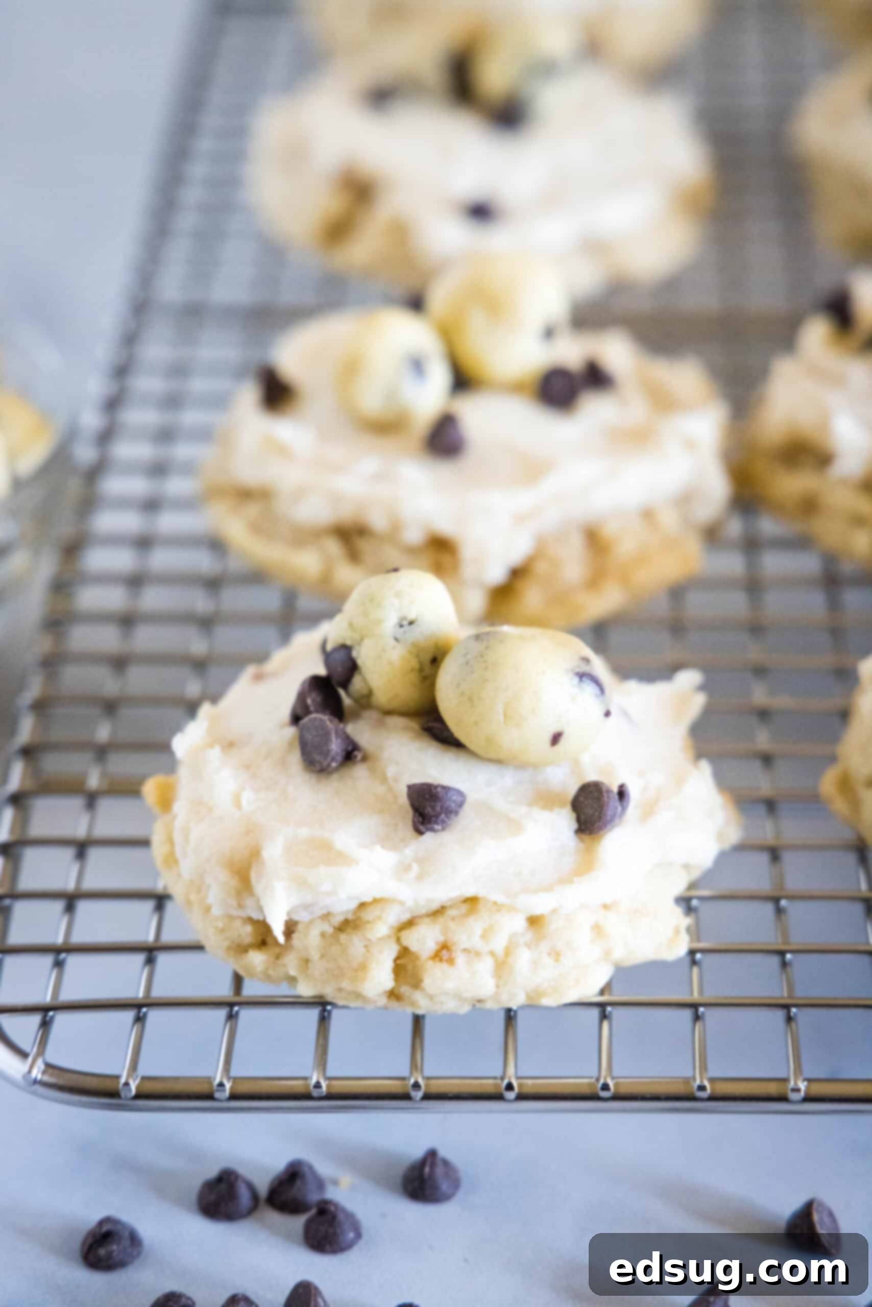 These cookie dough cookies are bakery-style sugar cookies topped with creamy cookie dough frosting, edible cookie dough, and chocolate chips! A close-up shot of several frosted cookie dough cookies, adorned with edible cookie dough and chocolate chips, presented on a wire cooling rack.