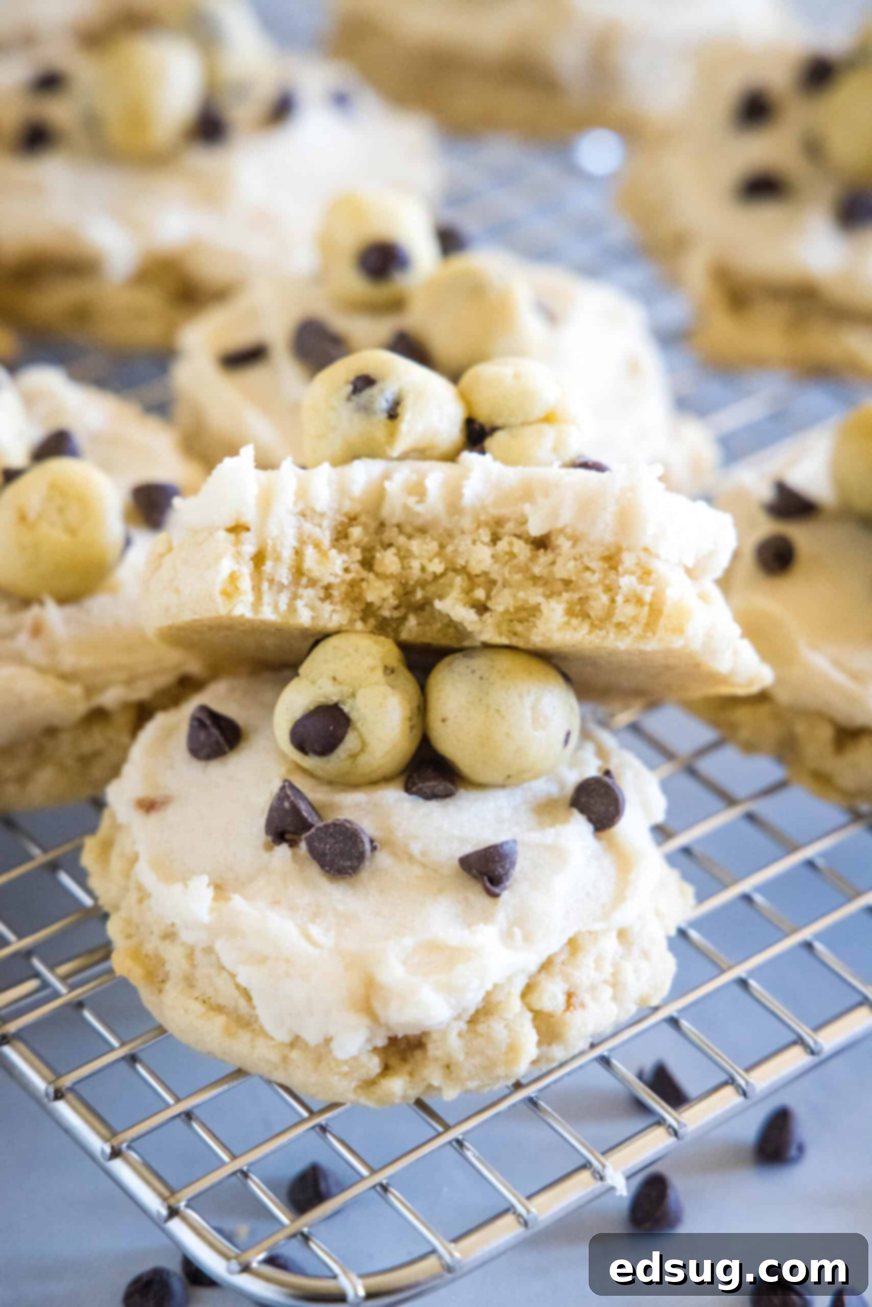 These cookie dough cookies are bakery-style sugar cookies topped with creamy cookie dough frosting, edible cookie dough, and chocolate chips! A close-up of frosted cookie dough cookies on a wire rack, with one cookie expertly broken in half to reveal its soft, chewy interior.
