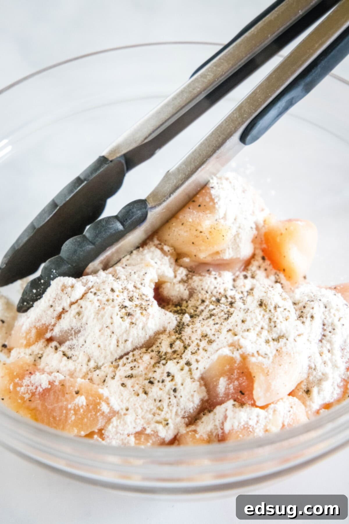 Flour being tossed with chicken pieces in a glass bowl using tongs.