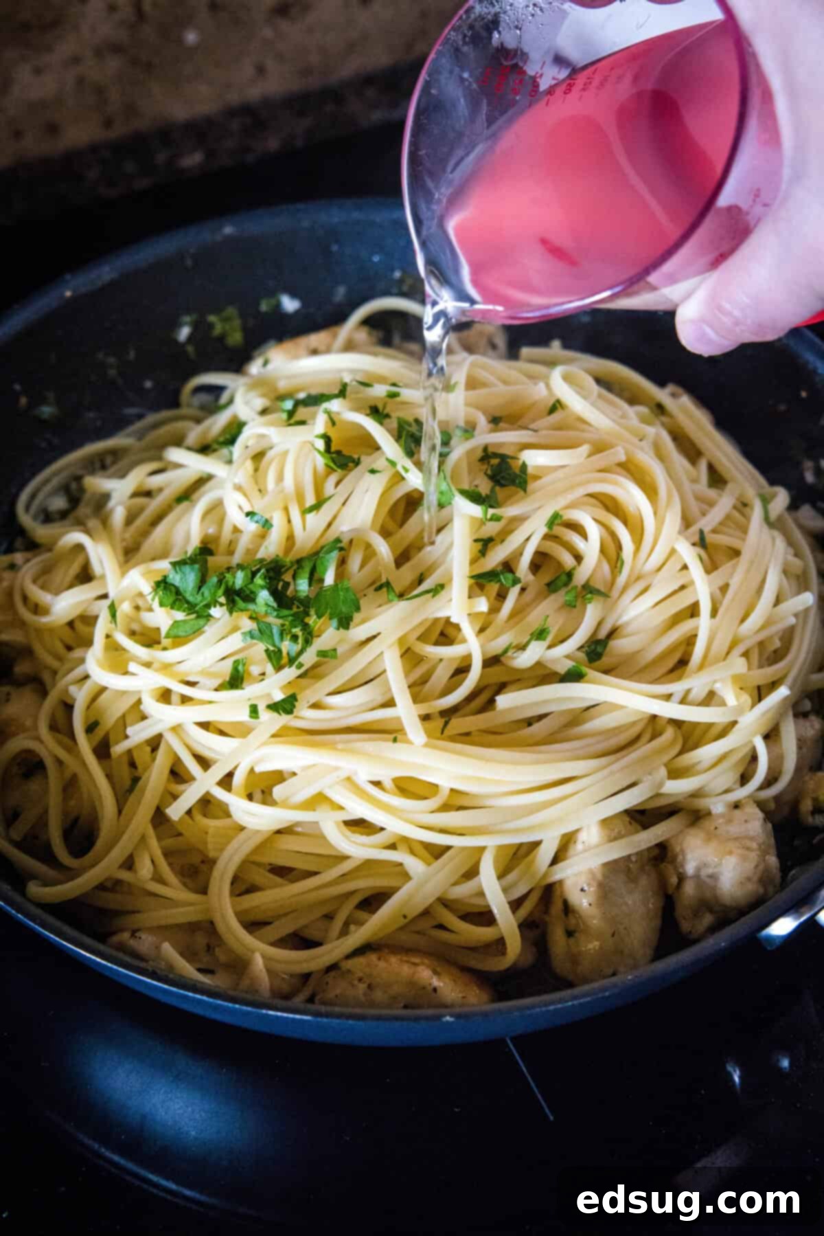 Cooked spaghetti and reserved pasta water being added to the chicken scampi in a skillet, then tossed.