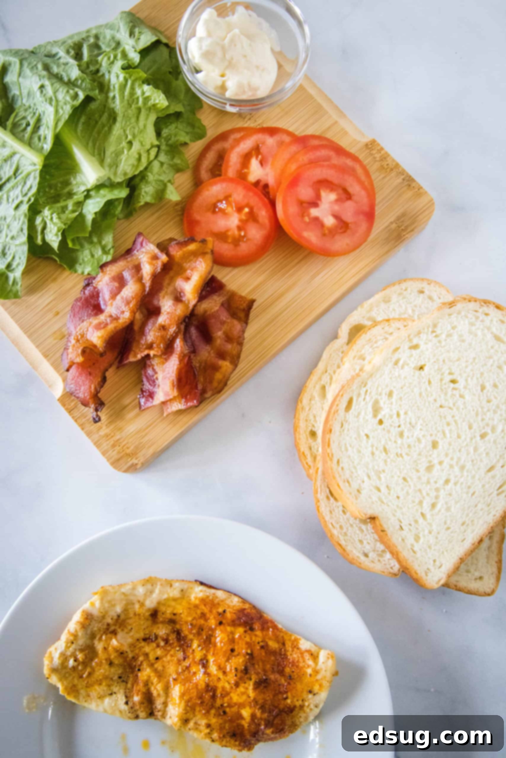 Various fresh ingredients laid out on a wooden surface, ready to be assembled into a chicken club sandwich, including bread, cooked chicken, bacon, lettuce, tomato, and mayonnaise.