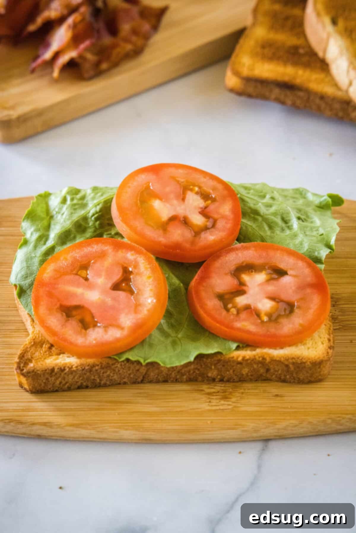 The initial layer of a chicken club sandwich being assembled, showing the first toasted bread slice topped with vibrant romaine lettuce and juicy tomato slices on a wooden cutting board.