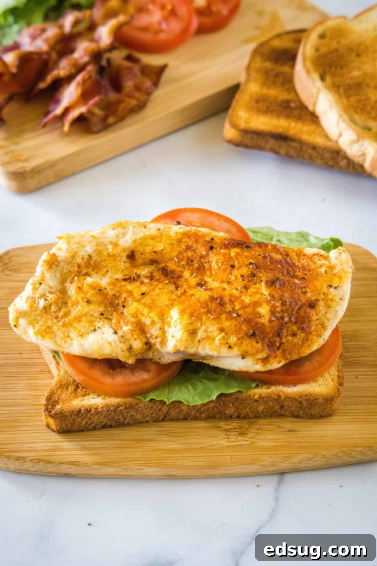 Building the first layer of a chicken club sandwich, featuring the toasted bread slice, lettuce, tomato, and a generous portion of perfectly seared or cooked chicken pieces.